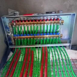 Rozdzielacz ogrzewania podłogowego w metalowej szafce, z widocznymi numerowanymi zaworami, zielonymi rurami i czerwonymi otulinami, zamontowany na folii z rastrem.