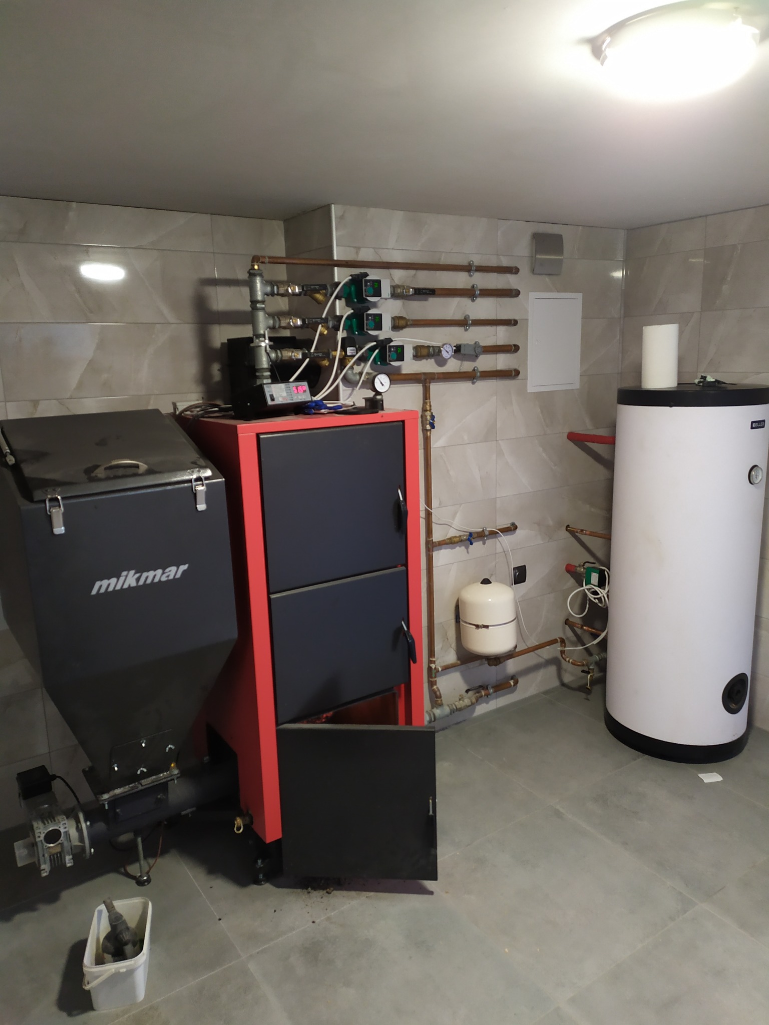 Kotłownia z kotłem na paliwo stałe Mikmar, zasobnikiem ciepłej wody użytkowej i instalacją centralnego ogrzewania z licznymi rurami miedzianymi i zaworami, widoczne również naczynie przeponowe...