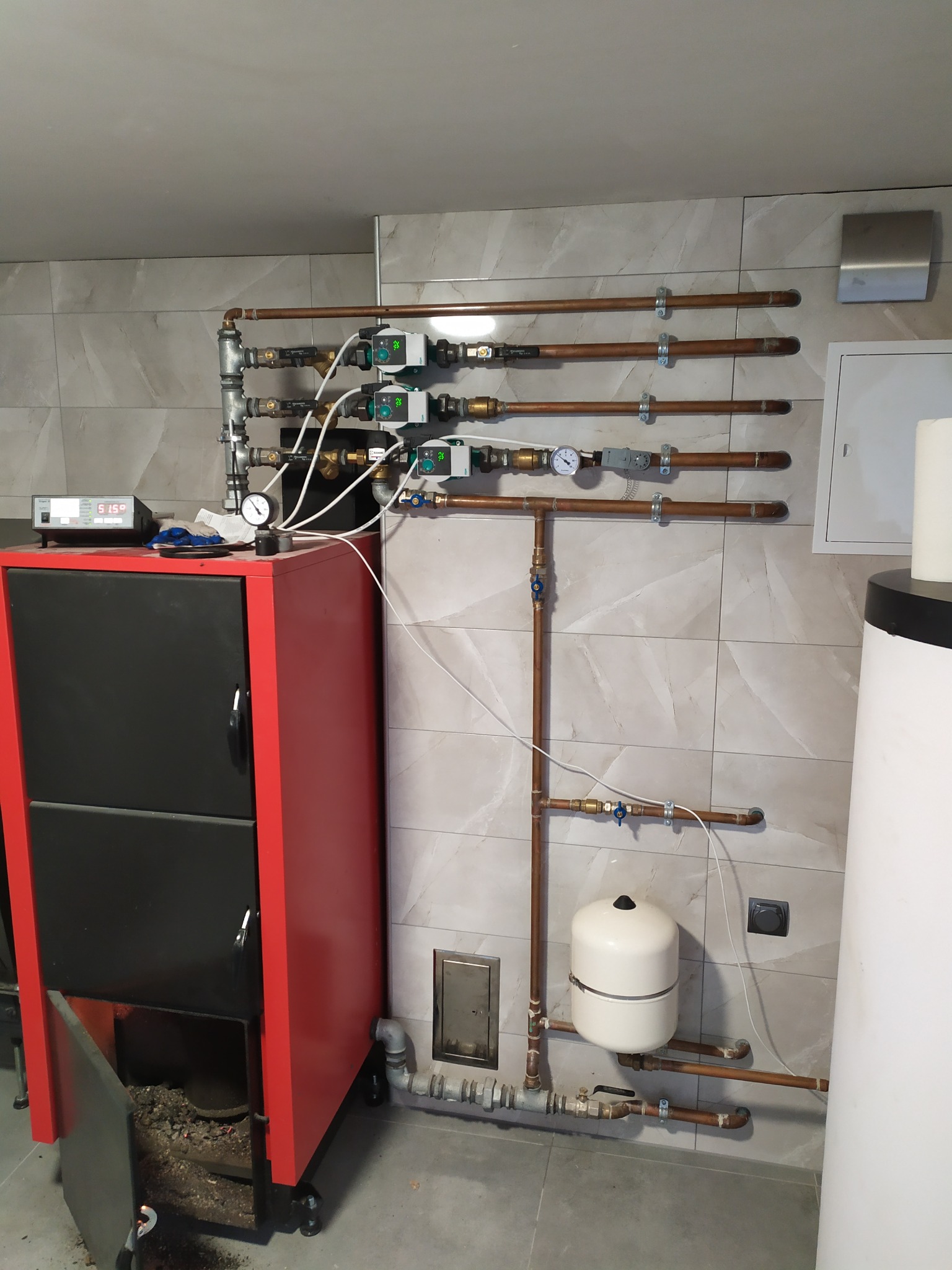 Czerwono-czarny piec z otwartymi drzwiczkami, widoczny popiół, połączony miedzianymi rurami z zaworami, manometrami i pompami obiegowymi na tle beżowych płytek ściennych.