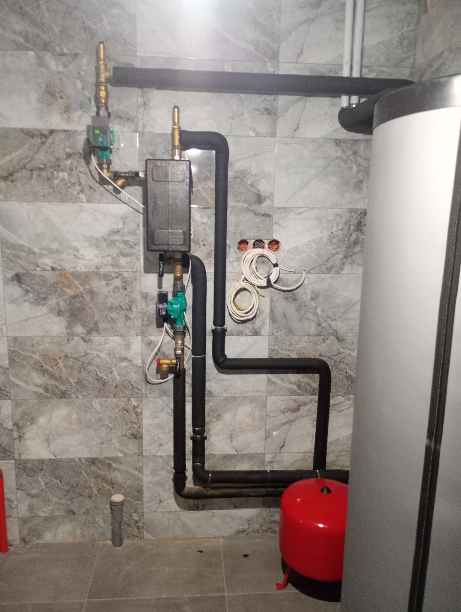 Nowoczesna kotłownia z szarymi płytkami imitującymi marmur, widoczne czarne rury instalacji grzewczej z zaworami, czerwony zbiornik wyrównawczy i biały zasobnik ciepłej wody, oraz wystające kable...