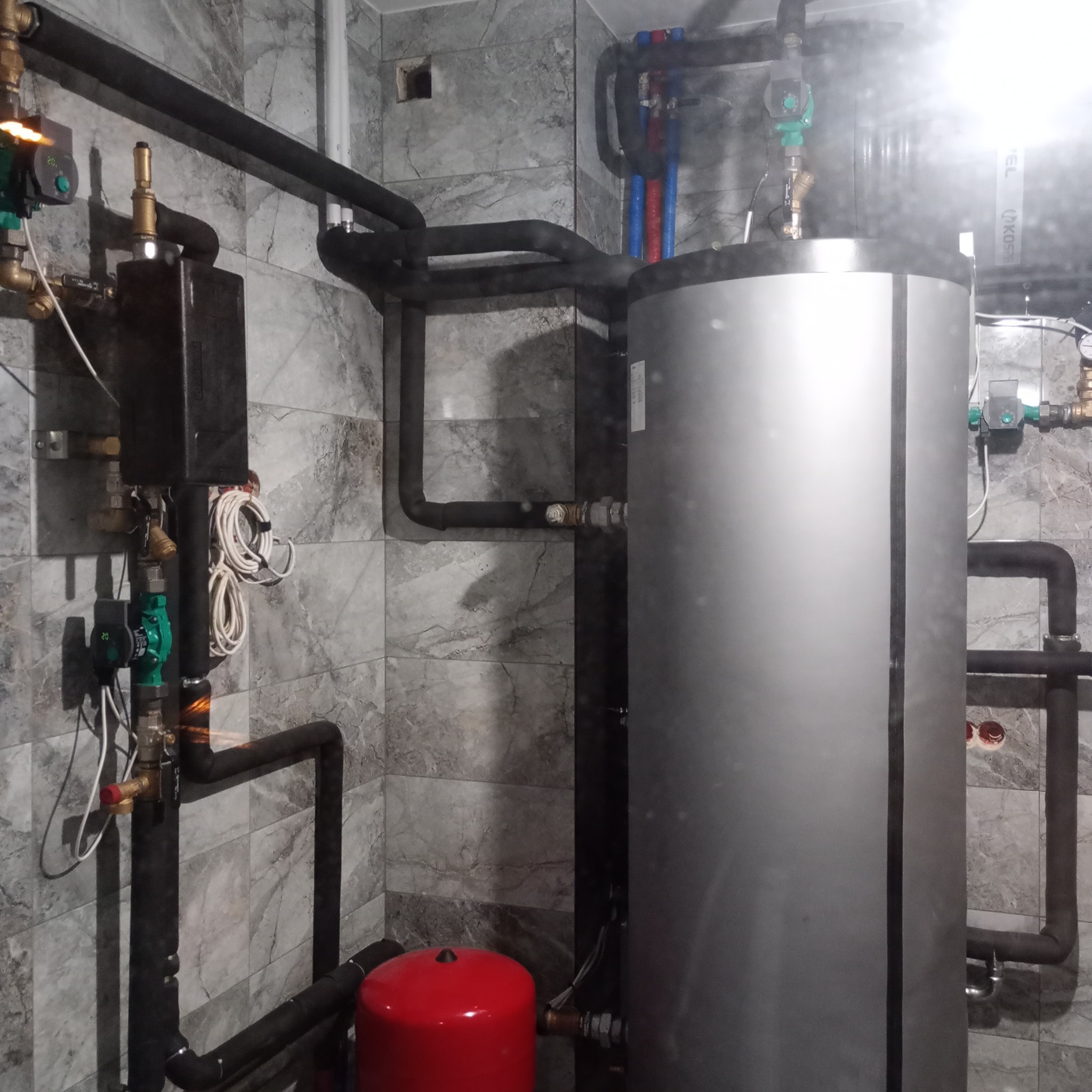 Szary bojler z czarnymi rurami izolowanymi termicznie, czerwony zbiornik wyrównawczy, zielone pompy obiegowe i szare płytki imitujące marmur na ścianie w kotłowni.