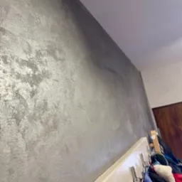 Ściana pokryta dekoracyjną, srebrzystą farbą strukturalną, widoczna z perspektywy dolnej, z fragmentem wieszaka na ubrania w tle.