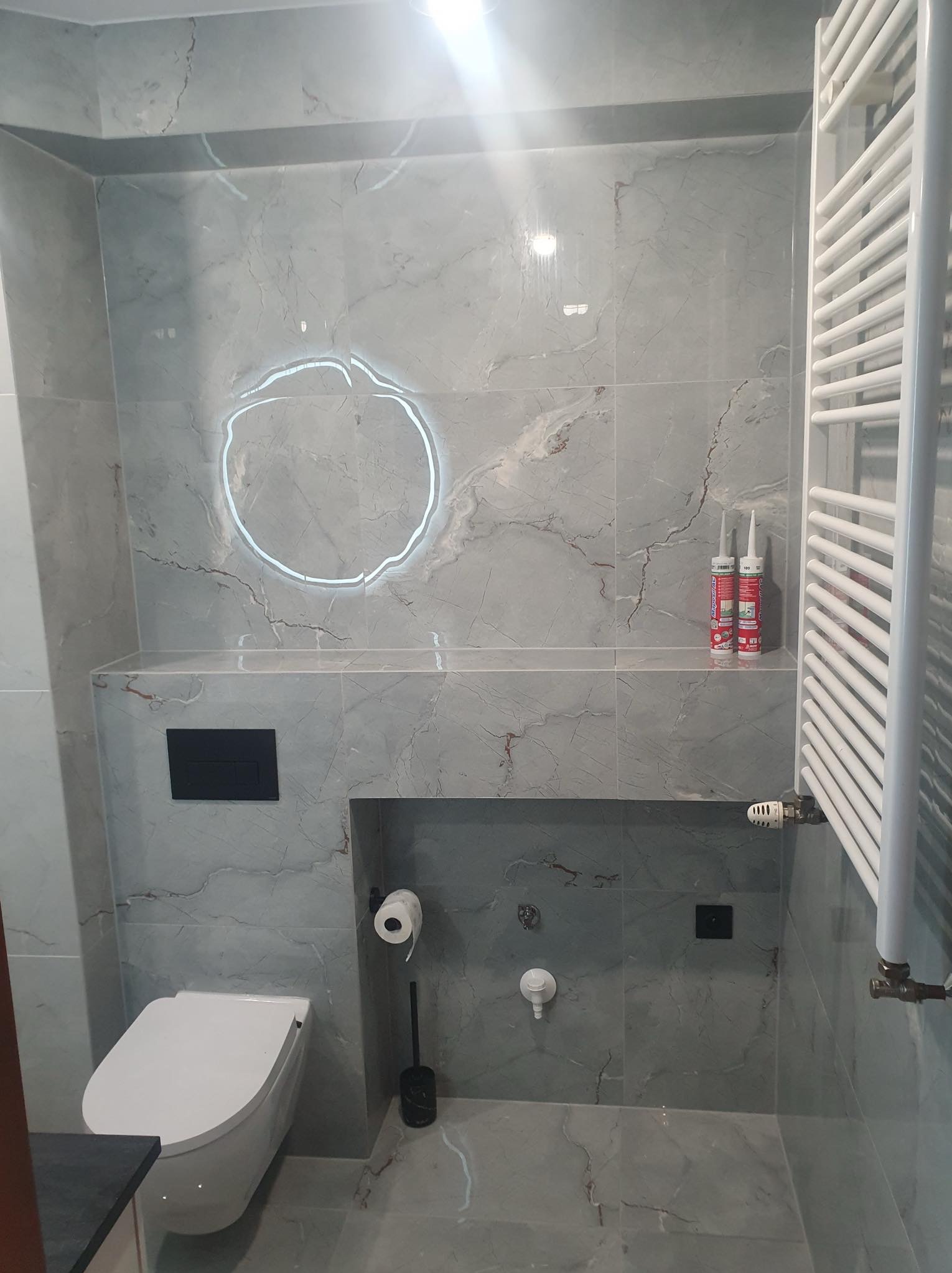 Nowoczesna łazienka z marmurowymi płytkami, podwieszaną toaletą, grzejnikiem drabinkowym i podświetlanym lustrem w kształcie okręgu, uzupełniona o tuby silikonu na półce.