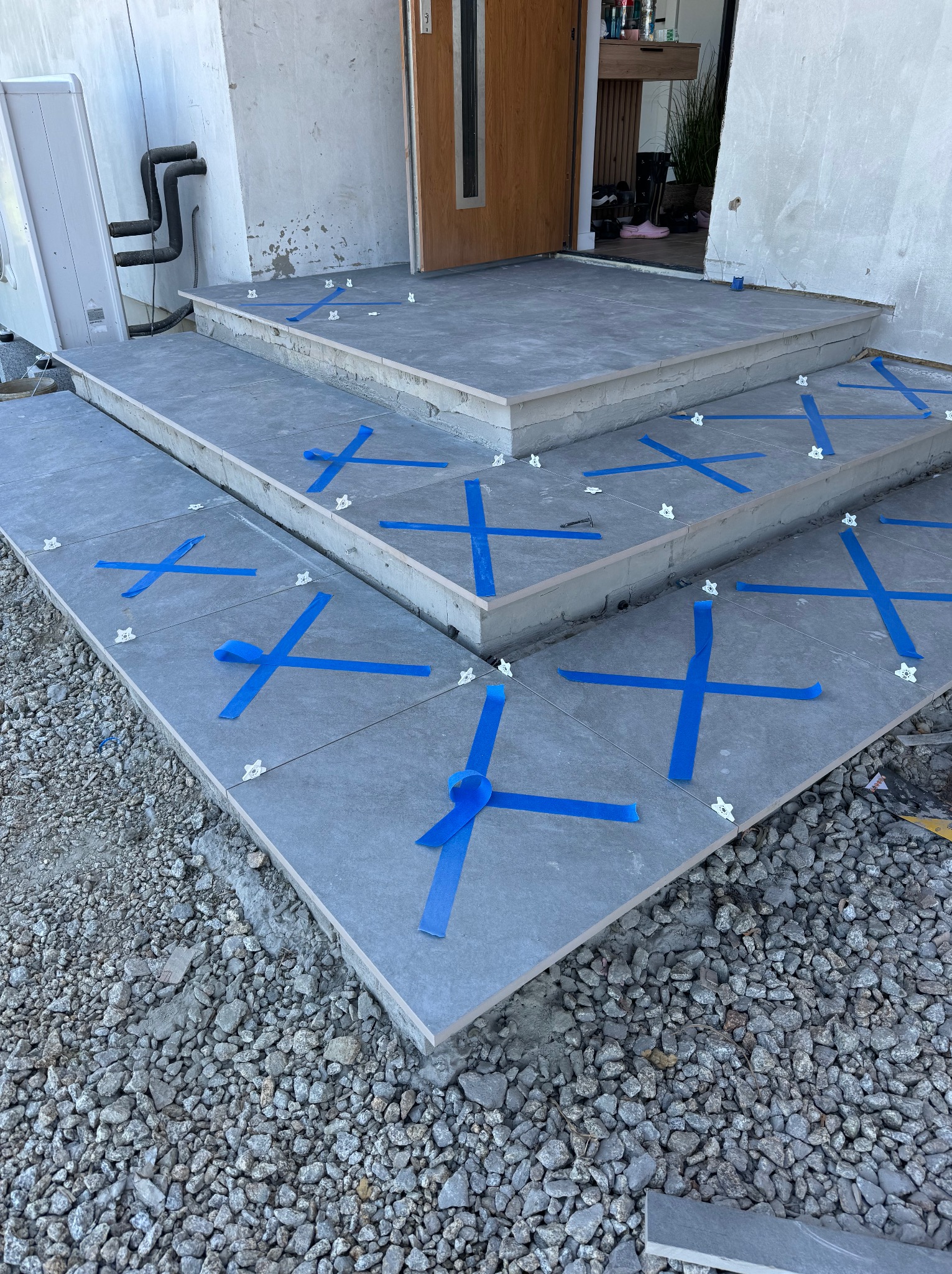 Układanie szarych płytek na schodach wejściowych, zabezpieczonych niebieską taśmą i plastikowymi krzyżykami dystansowymi, widok z góry na żwirowe podłoże.