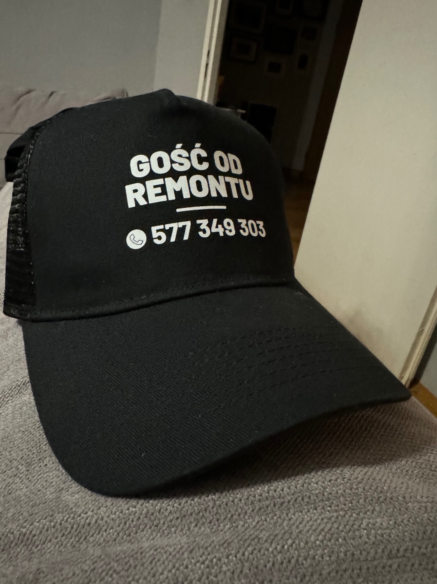 Czarna czapka z daszkiem z białym napisem 'GOŚĆ OD REMONTU' i numerem telefonu 577 349 303, leżąca na szarej kanapie.