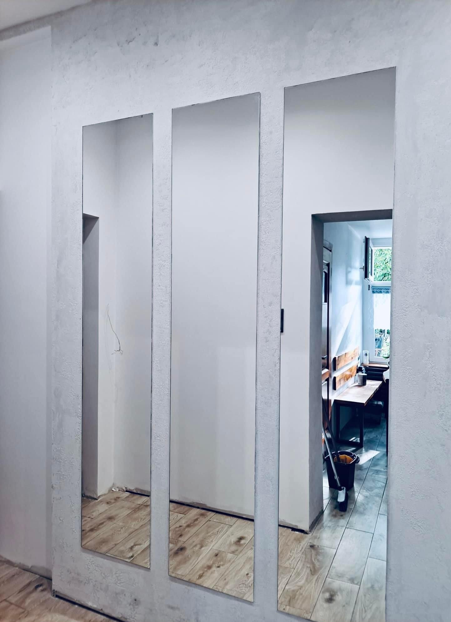 Trzy pionowe lustra bez ram osadzone w ścianie o szarej, chropowatej fakturze; widoczna podłoga z paneli imitujących drewno; w tle widok na pokój z drewnianym stołem i krzesłami, oraz wiadro i mop.