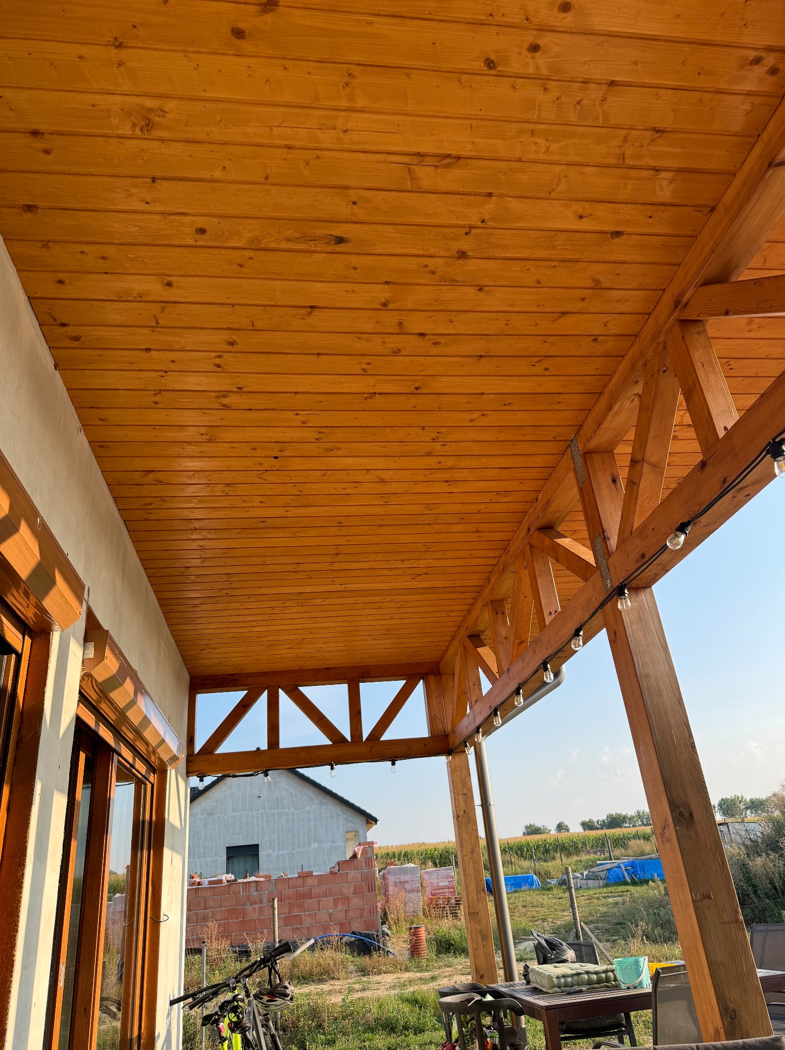 Zadaszony taras z drewnianą konstrukcją nośną i podbitką, oświetlony girlandą żarówek, z widokiem na budynek w trakcie budowy.