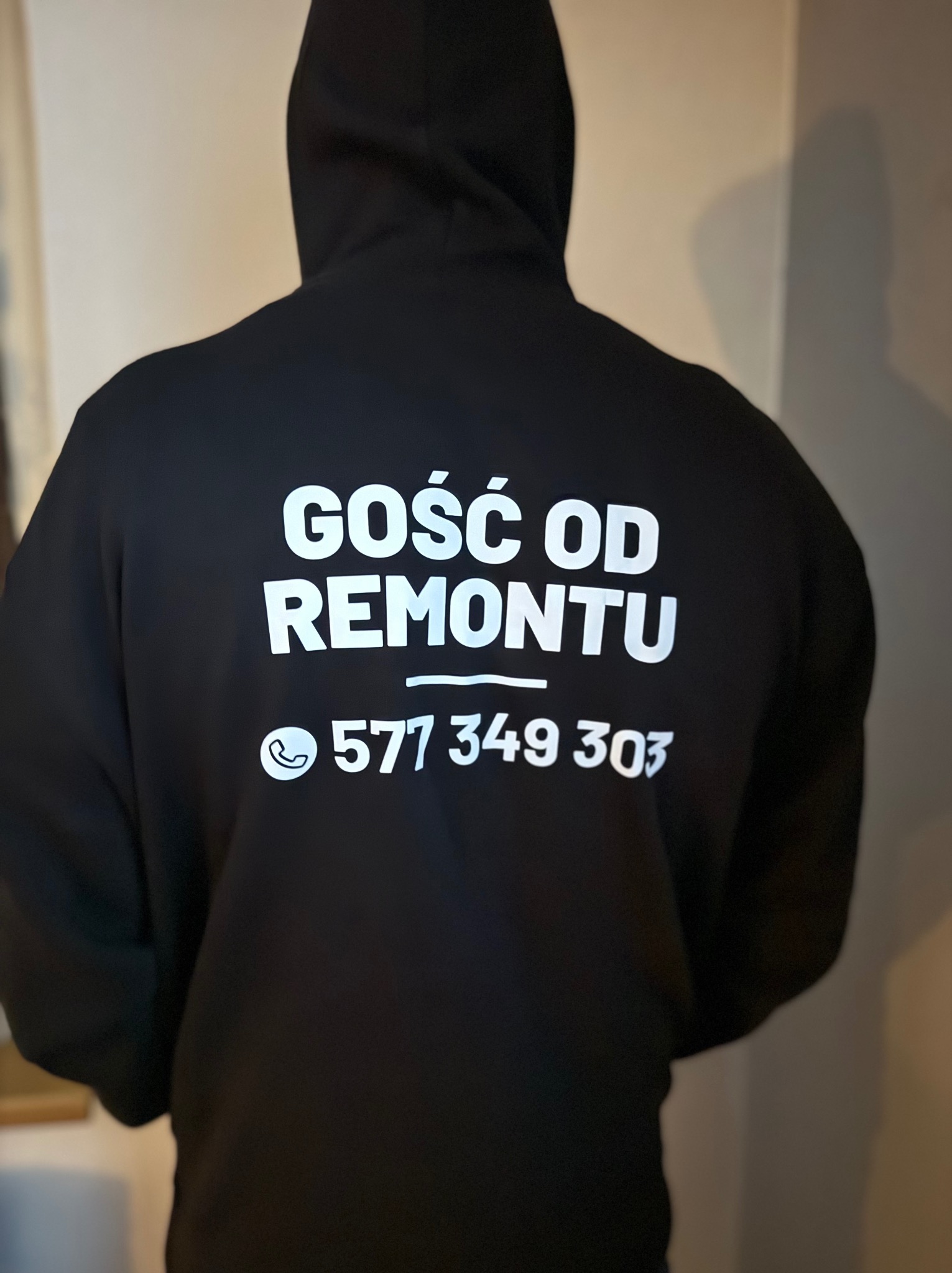 Czarna bluza z kapturem, na plecach biały napis 'GOŚĆ OD REMONTU' i numer telefonu 577 349 303, widok z tyłu.