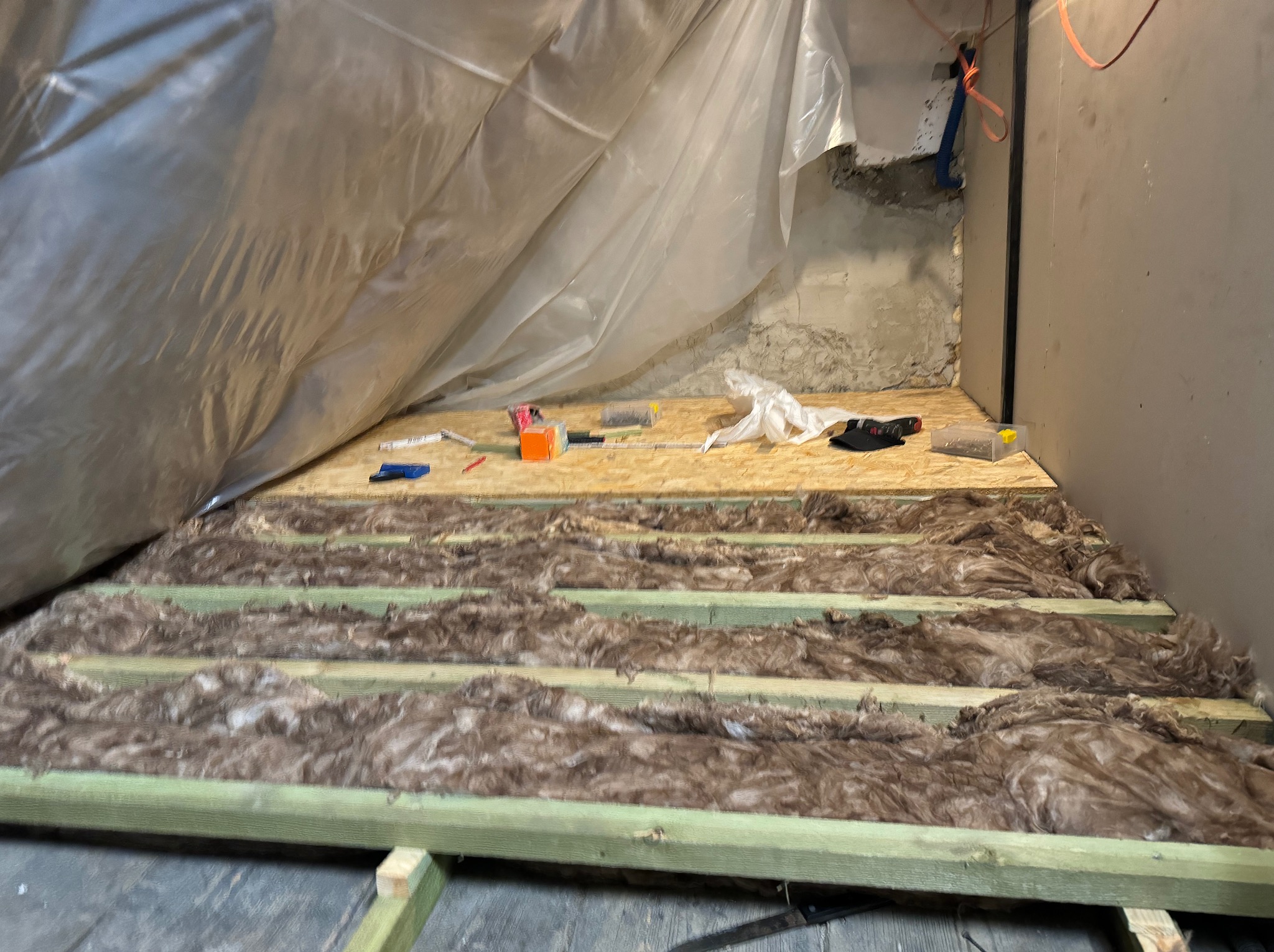 Izolacja poddasza wełną mineralną między legarami, częściowo zakryta folią, widoczne narzędzia i płyta OSB