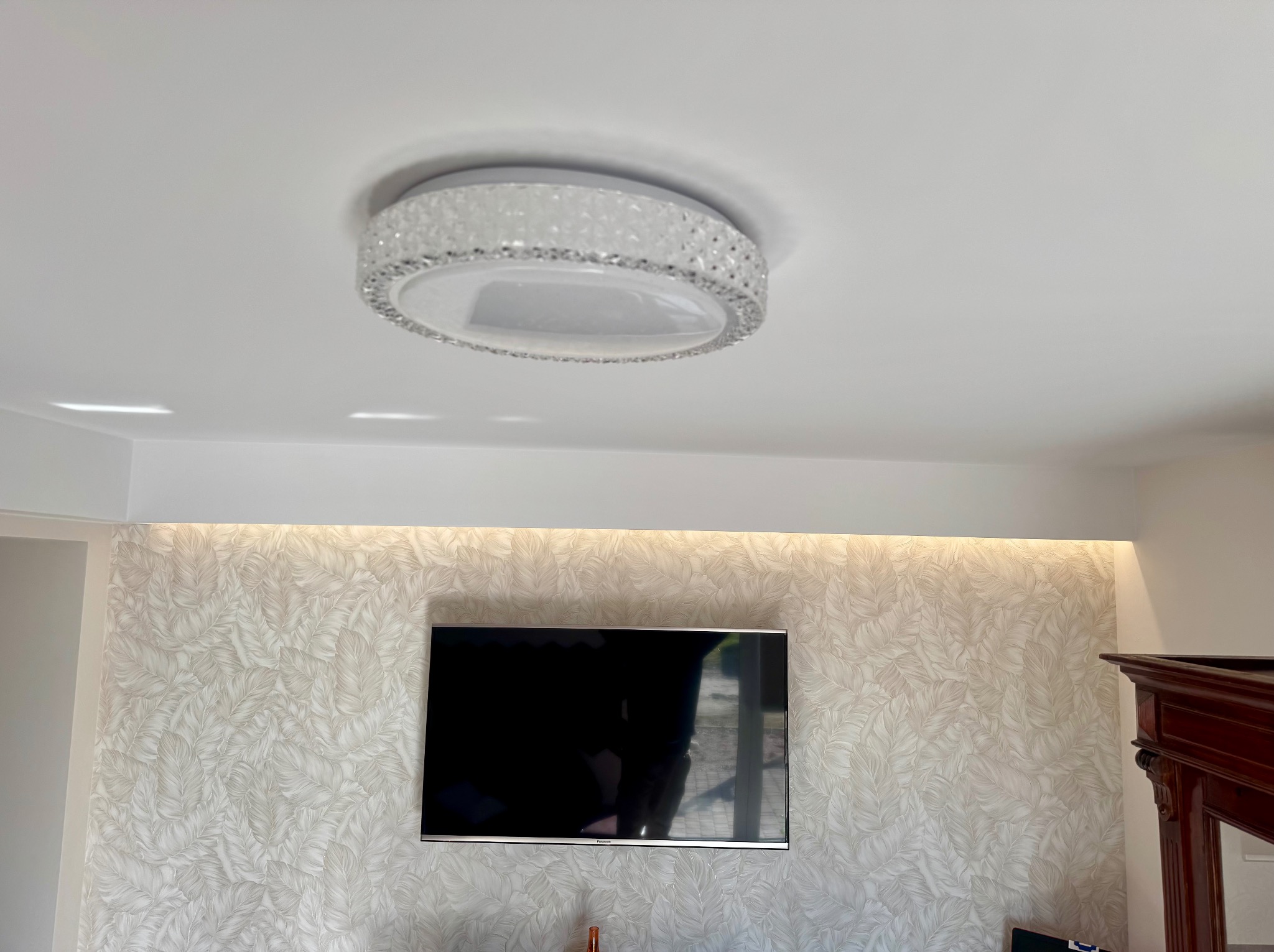 Kryształowy plafon sufitowy zamontowany w Świdnicy, podświetlona ściana z tapetą w liście, wiszący telewizor.