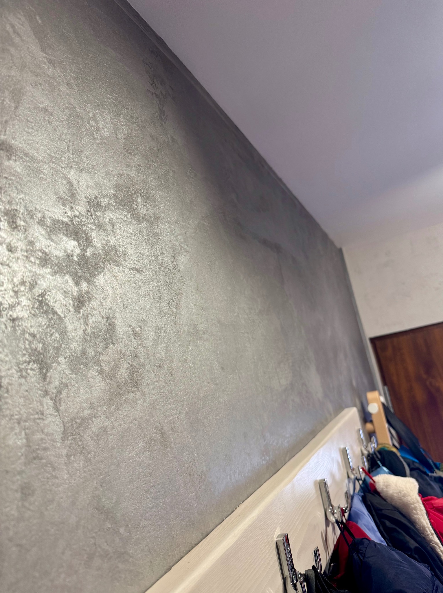 Ściana pokryta dekoracyjną, srebrzystą farbą strukturalną, widoczna z perspektywy dolnej, z fragmentem wieszaka na ubrania w tle.