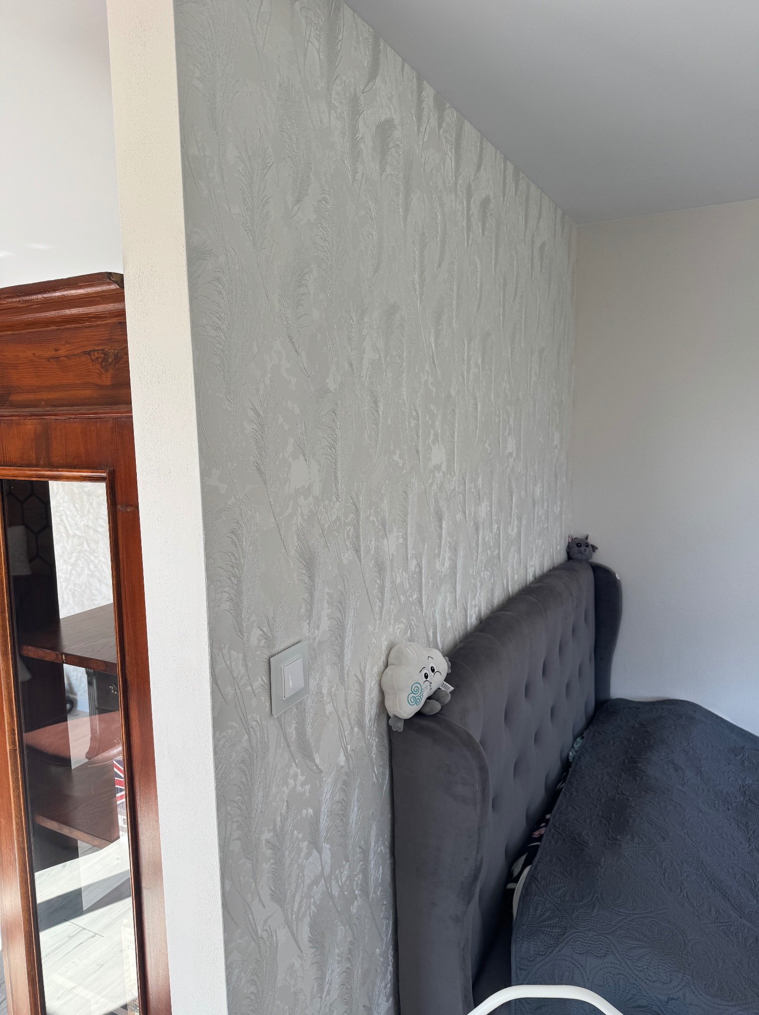 Ściana w sypialni z tapetą w delikatny, srebrzysty wzór piórkowy, obok drewniany regał z przeszkleniem, szary zagłówek łóżka z pluszowymi zabawkami.