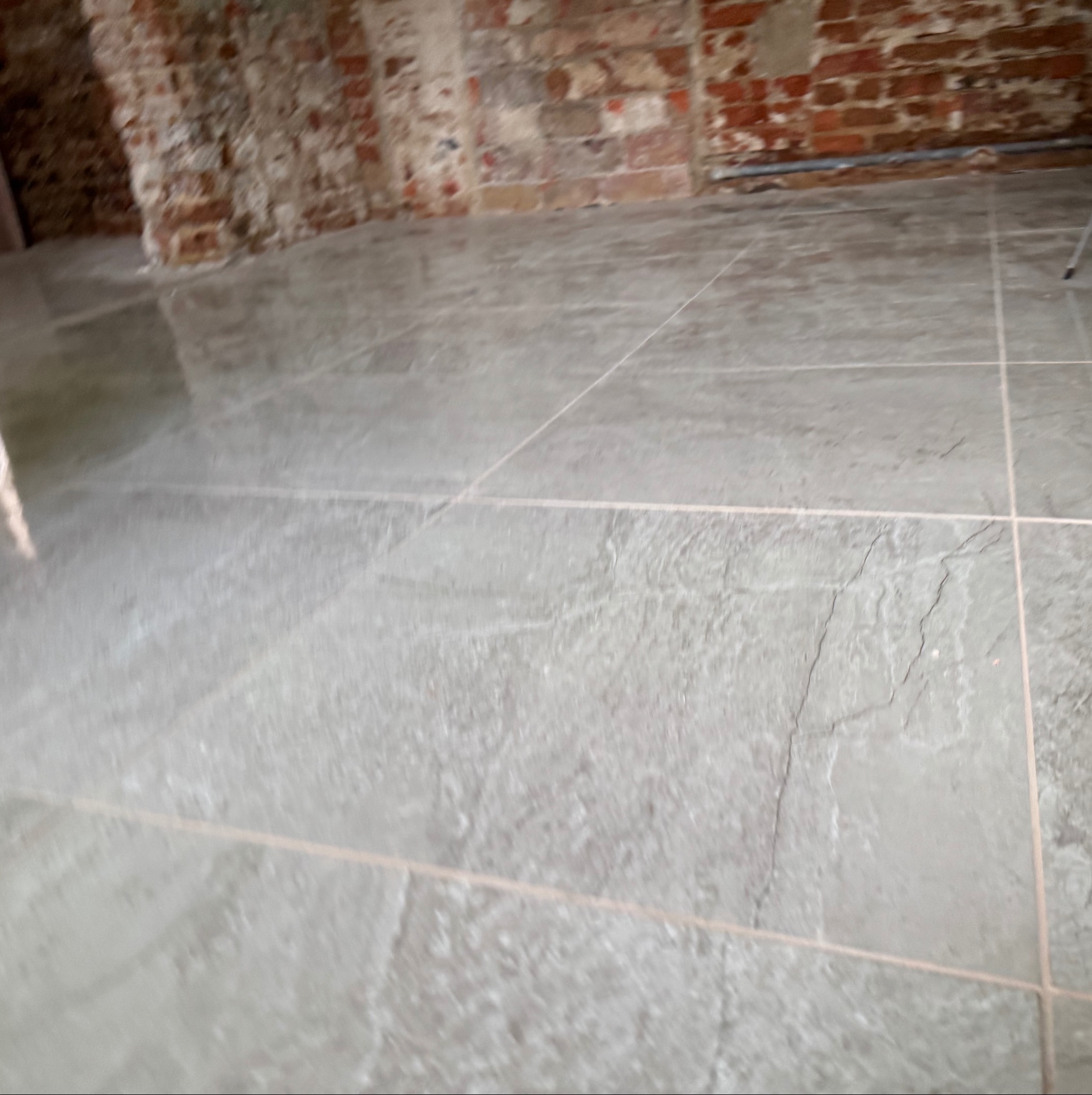 Świeżo położona, szara podłoga z płytek imitujących kamień, widoczne fugi, w tle fragmenty ściany z czerwonej cegły w surowym stanie.