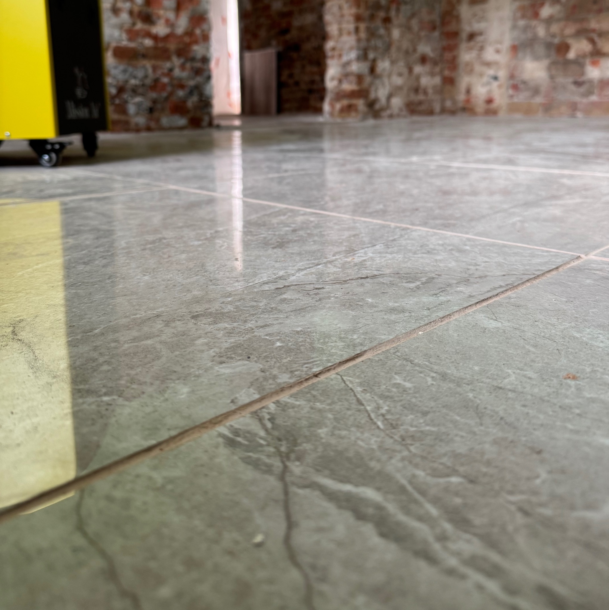 Szare płytki podłogowe z widoczną fugą, w tle ceglana ściana i żółty osuszacz powietrza.