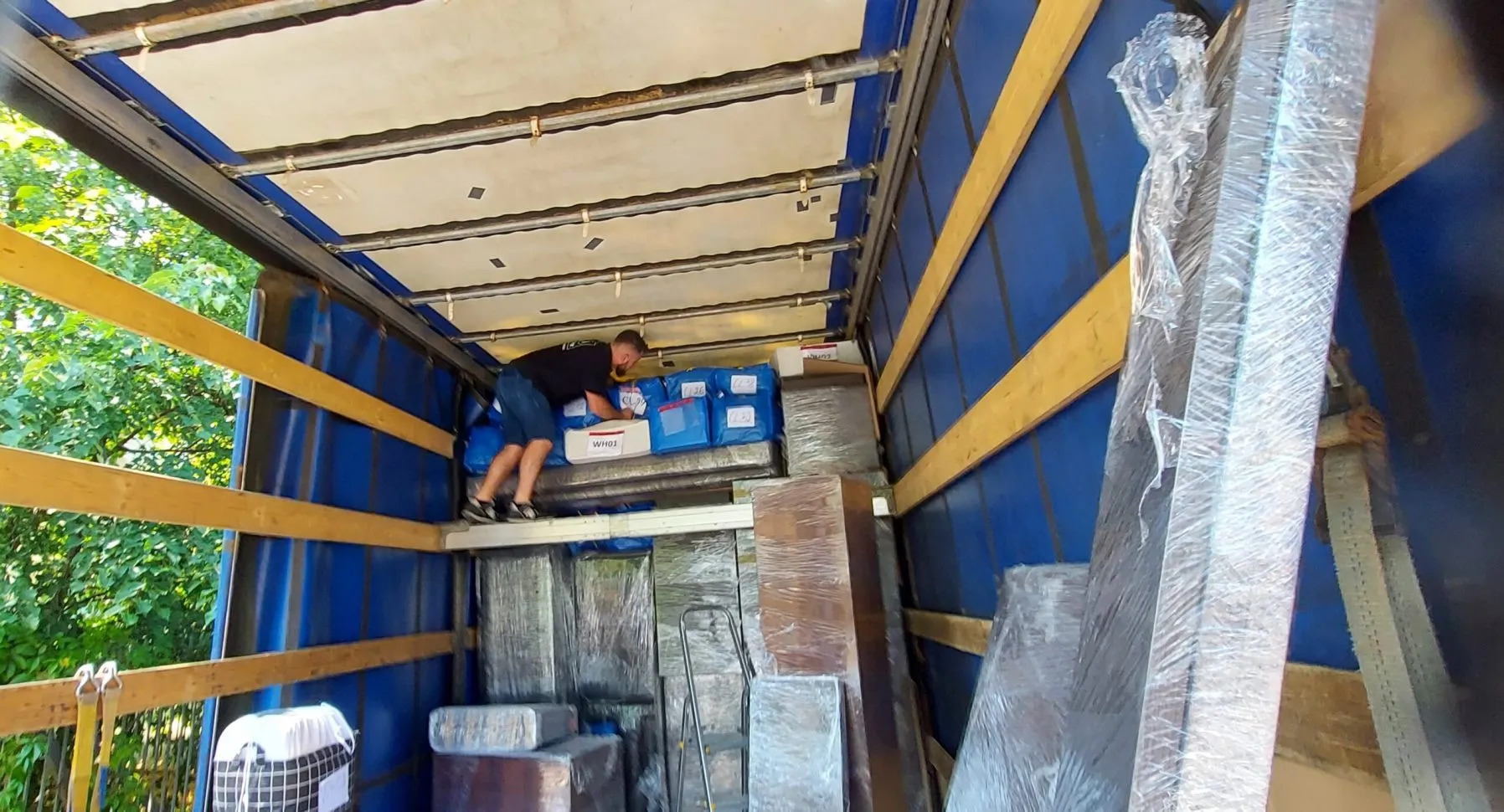 Wnętrze busa transportowego: mężczyzna układa zapakowane paczki i meble, zabezpieczone folią stretch, gotowe do przewozu. Widoczne niebieskie ściany i drewniane wzmocnienia.