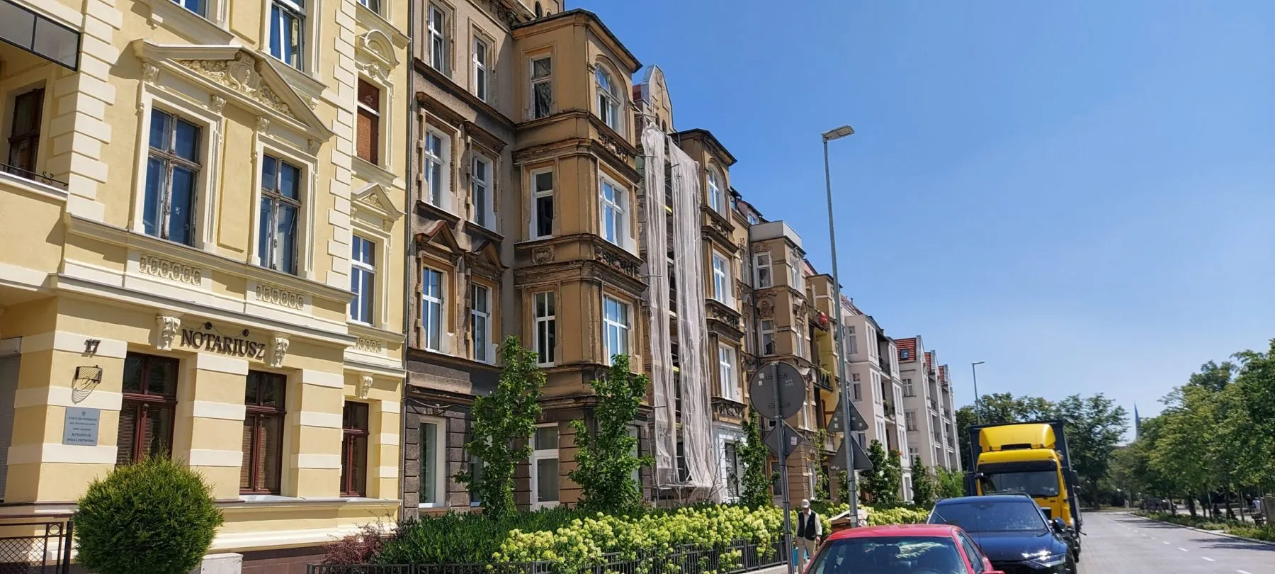 Ulica z kamienicami w różnym stanie, jedna w trakcie remontu z siatką ochronną. Na pierwszym planie zaparkowane auta i zieleń. W tle widoczny żółty samochód dostawczy.