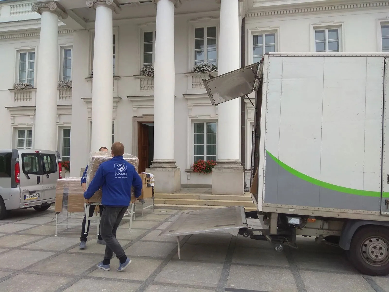 Przeprowadzka na tle klasycystycznego budynku z kolumnami. Pracownicy w niebieskich bluzach wnoszą zapakowane meble do ciężarówki z rampą. Widoczny bus i kostka brukowa.