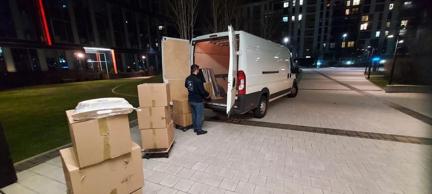 Wieczorny załadunek kartonów i mebli do białego busa na tle nowoczesnego budynku. Pracownik w ciemnej bluzie wkłada elementy do wnętrza pojazdu.