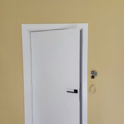 Oleksandr Turchynskyi - Ściana w kolorze piaskowym z zamontowanymi, białymi drzwiami z czarną klamką; widoczne niedokończone gniazdko elektryczne i kabel na haczyku.