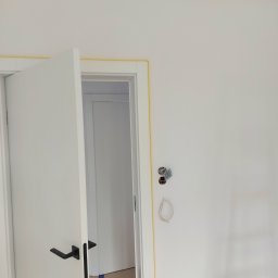 Oleksandr Turchynskyi - Świeżo pomalowana biała ściana z widoczną puszką elektryczną, zabezpieczona żółtą taśmą malarską przy suficie i ościeżnicy drzwi z czarną klamką, w tle widoczne wnętrze innego pomieszczenia.