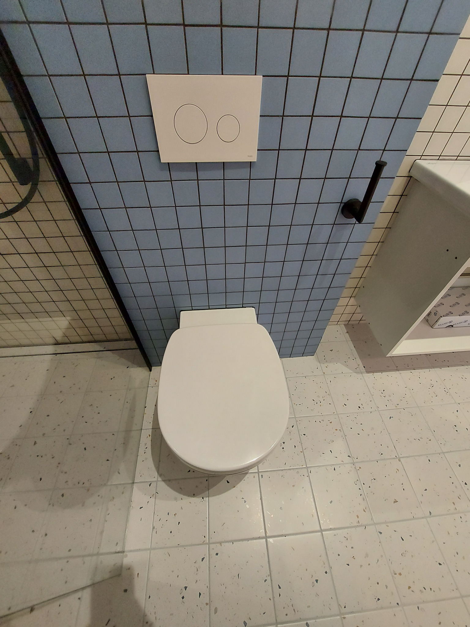 Gotowa łazienka we Wrocławiu: widok z góry na białą toaletę, błękitne kafelki z ciemną fugą, czarny uchwyt na papier toaletowy i szafkę z umywalką.