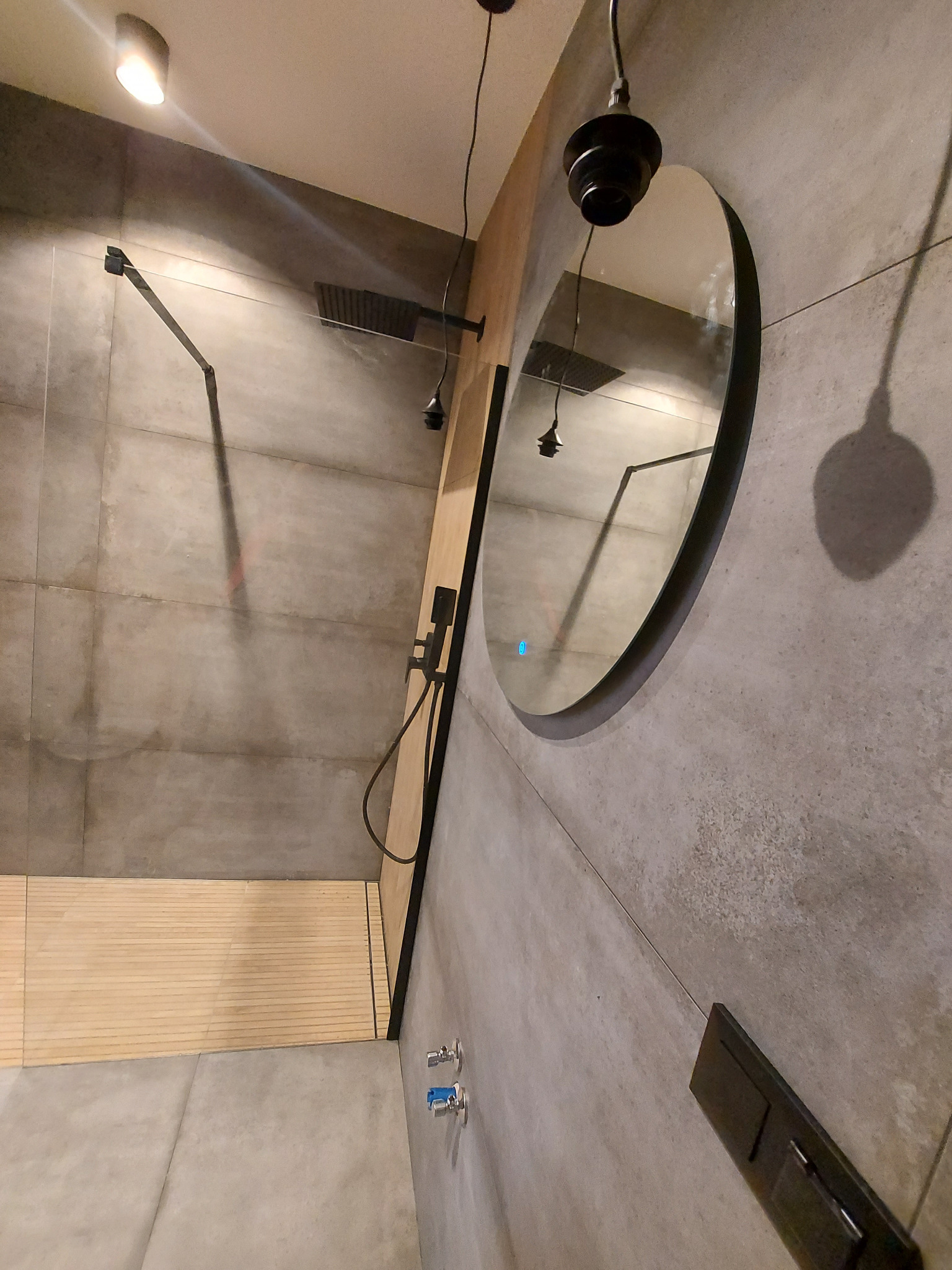 Nowoczesna łazienka z szarymi płytkami, deszczownicą, okrągłym lustrem z podświetleniem LED i drewnianym podestem prysznicowym.