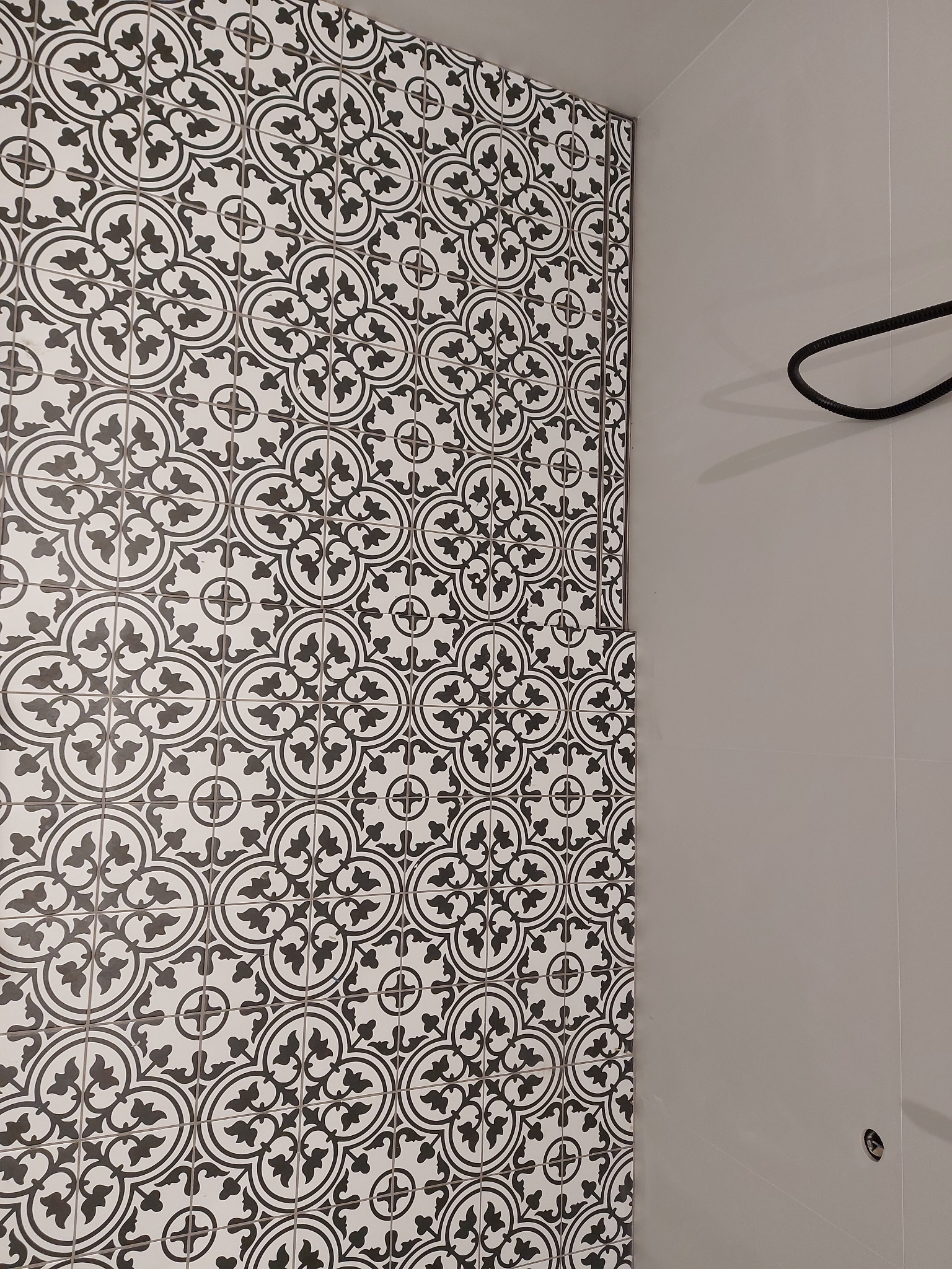 Fragment wykończonej łazienki z biało-czarną wzorzystą ścianą z płytek ceramicznych, sąsiadującą z gładką, szarą powierzchnią i fragmentem czarnego wieszaka.