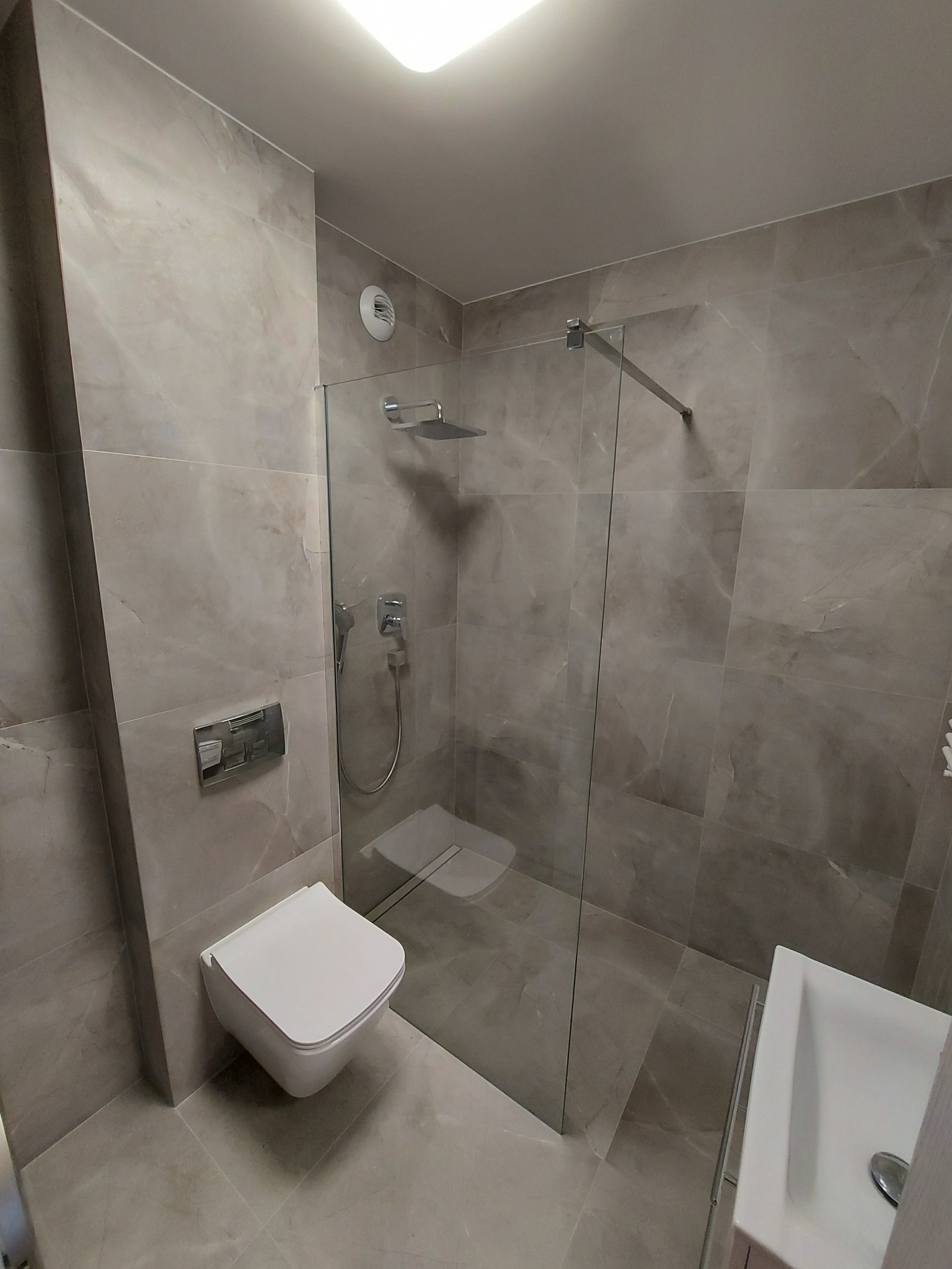 Nowoczesna łazienka z szarymi płytkami imitującymi marmur, kabina prysznicowa typu walk-in ze szklaną ścianką, podwieszana toaleta i umywalka nablatowa, oświetlenie sufitowe.