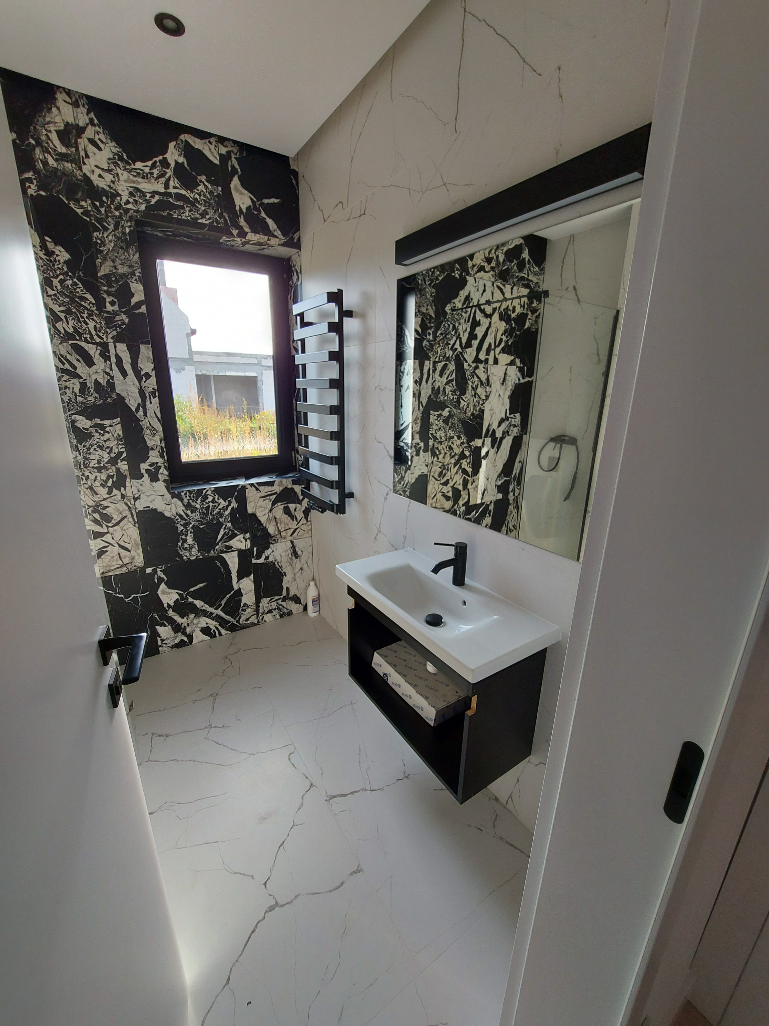 Wykończona łazienka we Wrocławiu z czarno-białymi marmurkowymi płytkami na ścianach i podłodze, z czarną armaturą i grzejnikiem drabinkowym oraz oknem z widokiem na budowę.
