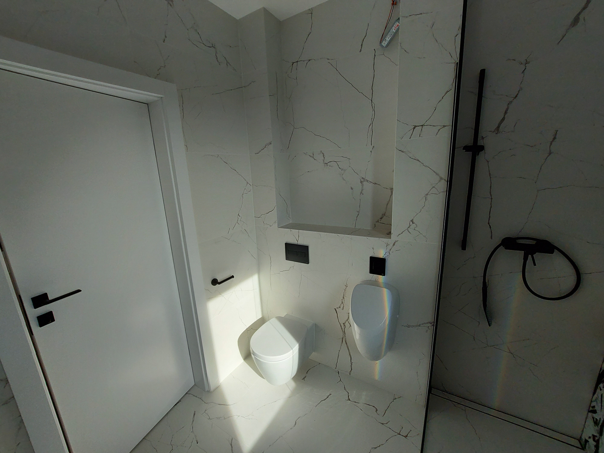 Nowoczesna łazienka z białymi płytkami imitującymi marmur, wiszącą toaletą i pisuar oraz czarnymi akcentami w postaci armatury i uchwytów, widoczne przewody elektryczne w górnym rogu.