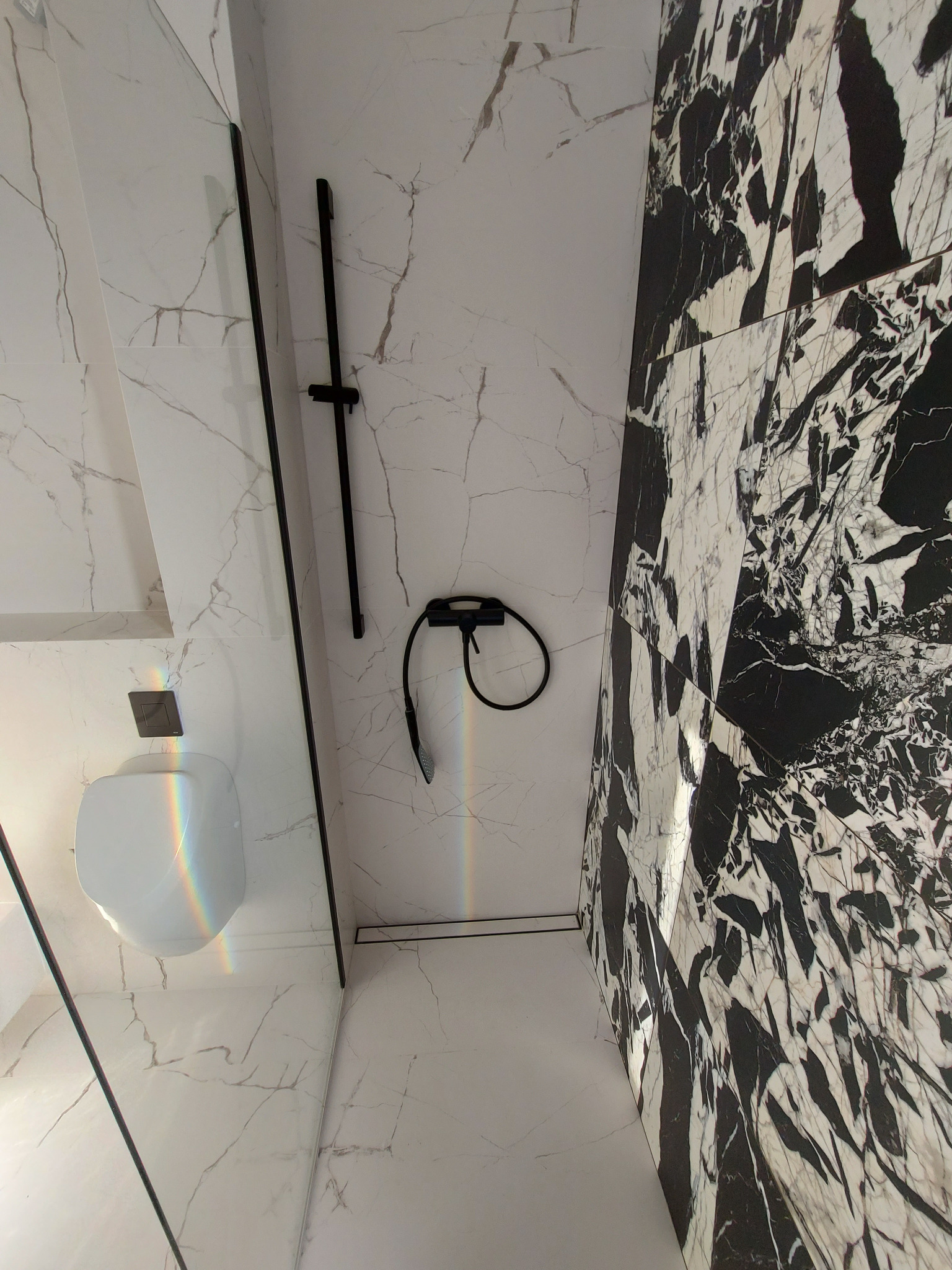 Nowoczesna łazienka z kabiną prysznicową typu walk-in, wykończona białymi płytkami imitującymi marmur z szarymi żyłami oraz czarnymi płytkami marmurowymi z białymi żyłami, czarna armatura, widoczny...