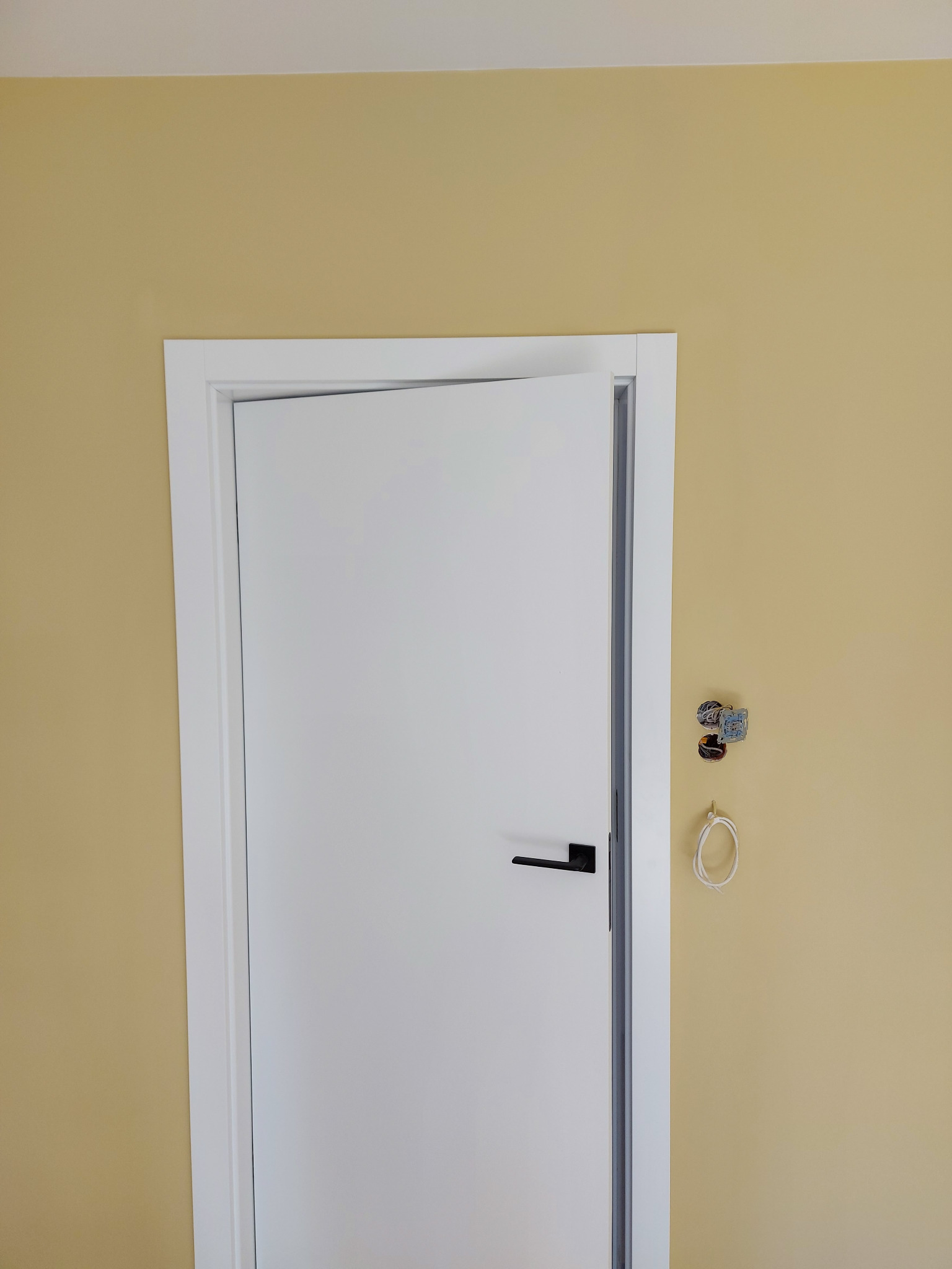Ściana w kolorze piaskowym z zamontowanymi, białymi drzwiami z czarną klamką; widoczne niedokończone gniazdko elektryczne i kabel na haczyku.