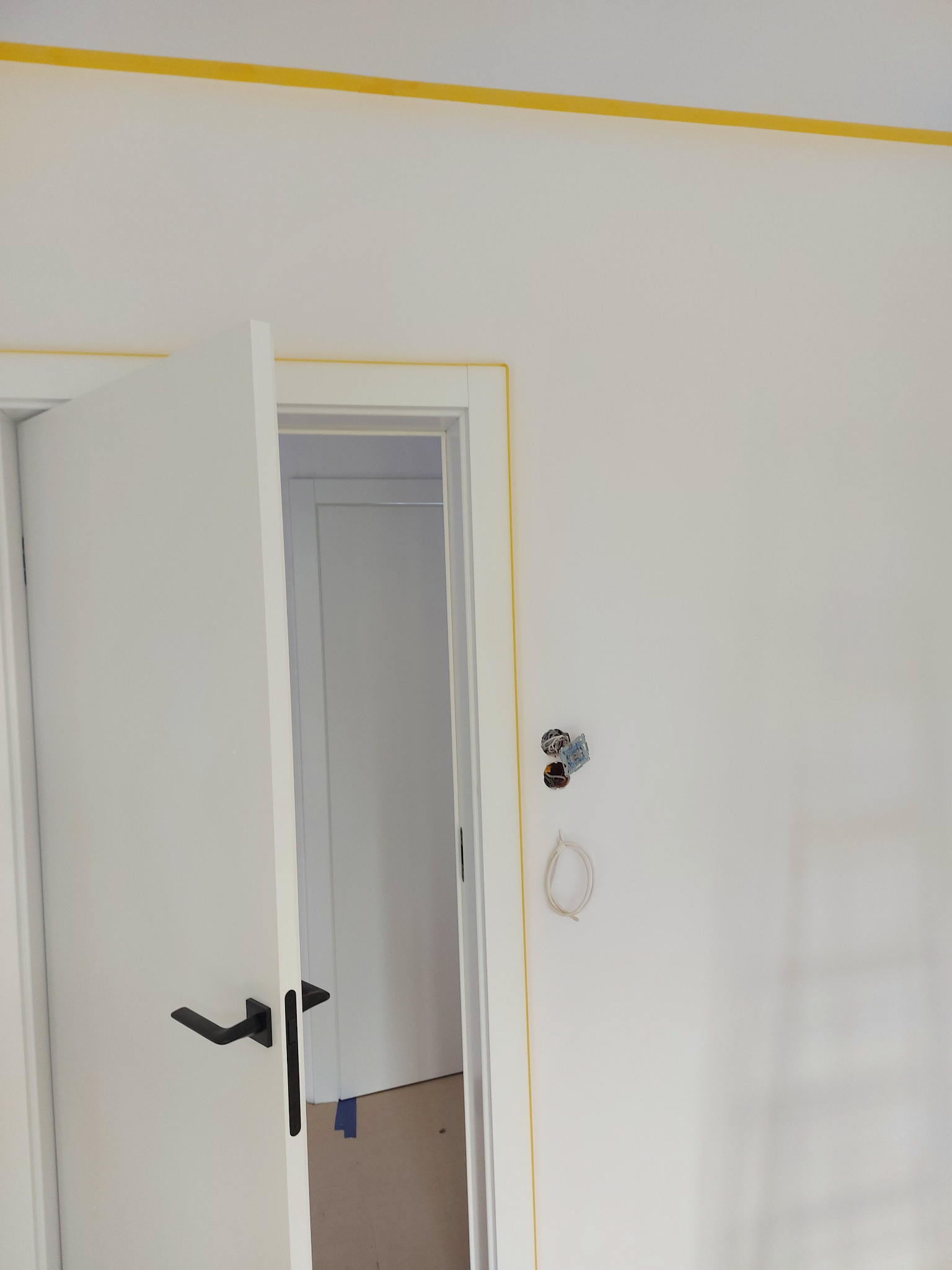 Świeżo pomalowana biała ściana z widoczną puszką elektryczną, zabezpieczona żółtą taśmą malarską przy suficie i ościeżnicy drzwi z czarną klamką, w tle widoczne wnętrze innego pomieszczenia.