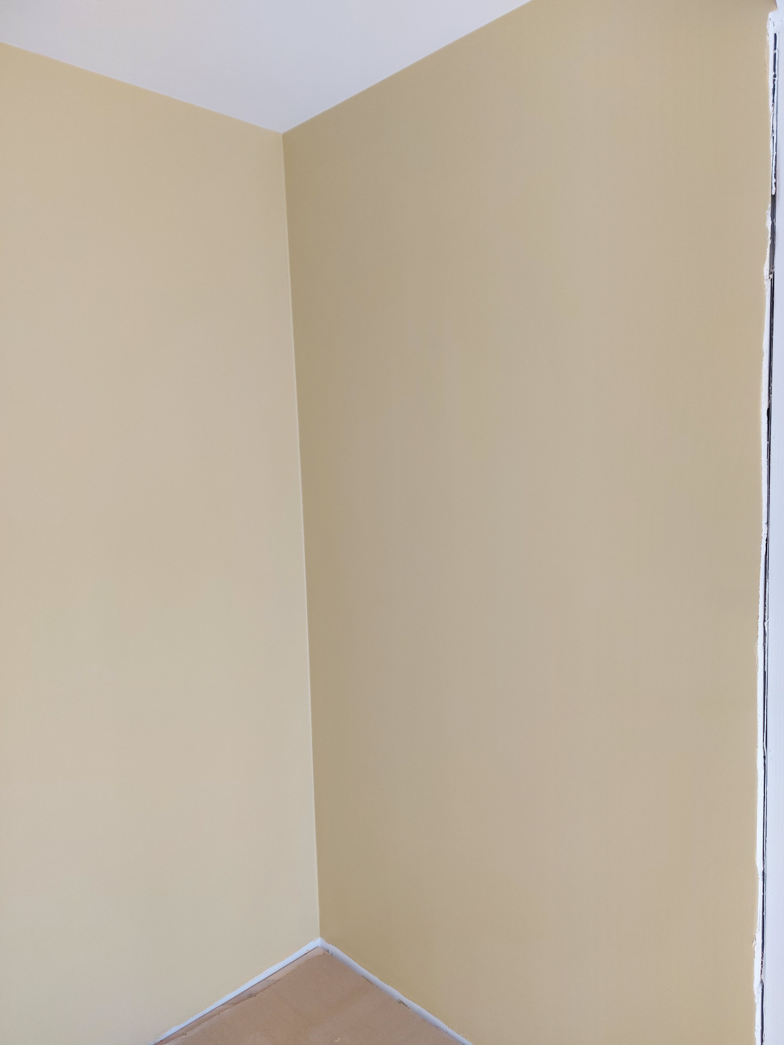 Świeżo pomalowany narożnik pokoju na jasny, ciepły odcień beżu, z widocznymi zabezpieczeniami podłogi i listew przypodłogowych.