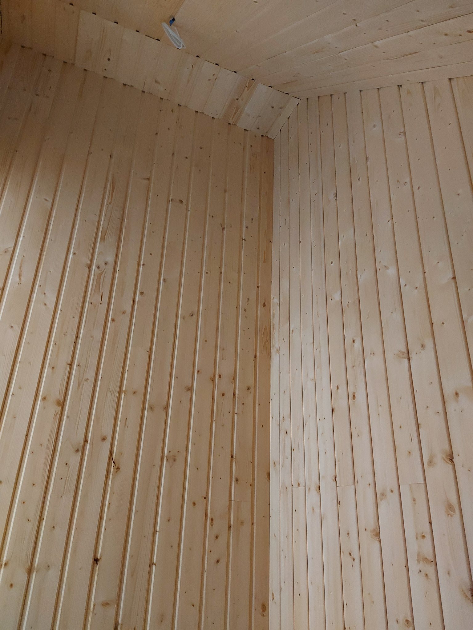 Jasne, pionowo ułożone deski boazerii pokrywają ściany i sufit pomieszczenia, tworząc kąt w rogu, z widocznym fragmentem instalacji elektrycznej na suficie.