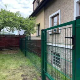 Zielone ogrodzenie panelowe z bramką na tle budynku z oknami i drzewa. Widok z zewnątrz, na trawniku.