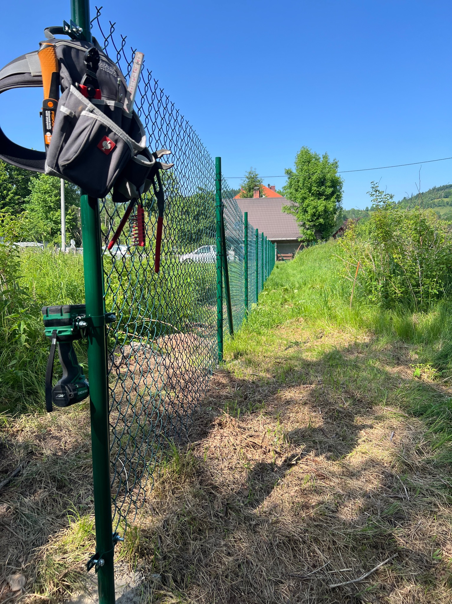 Fragment montażu zielonej siatki ogrodzeniowej z narzędziami przy słupku. W tle trawa, drzewa i dom. Widoczny pas narzędziowy.