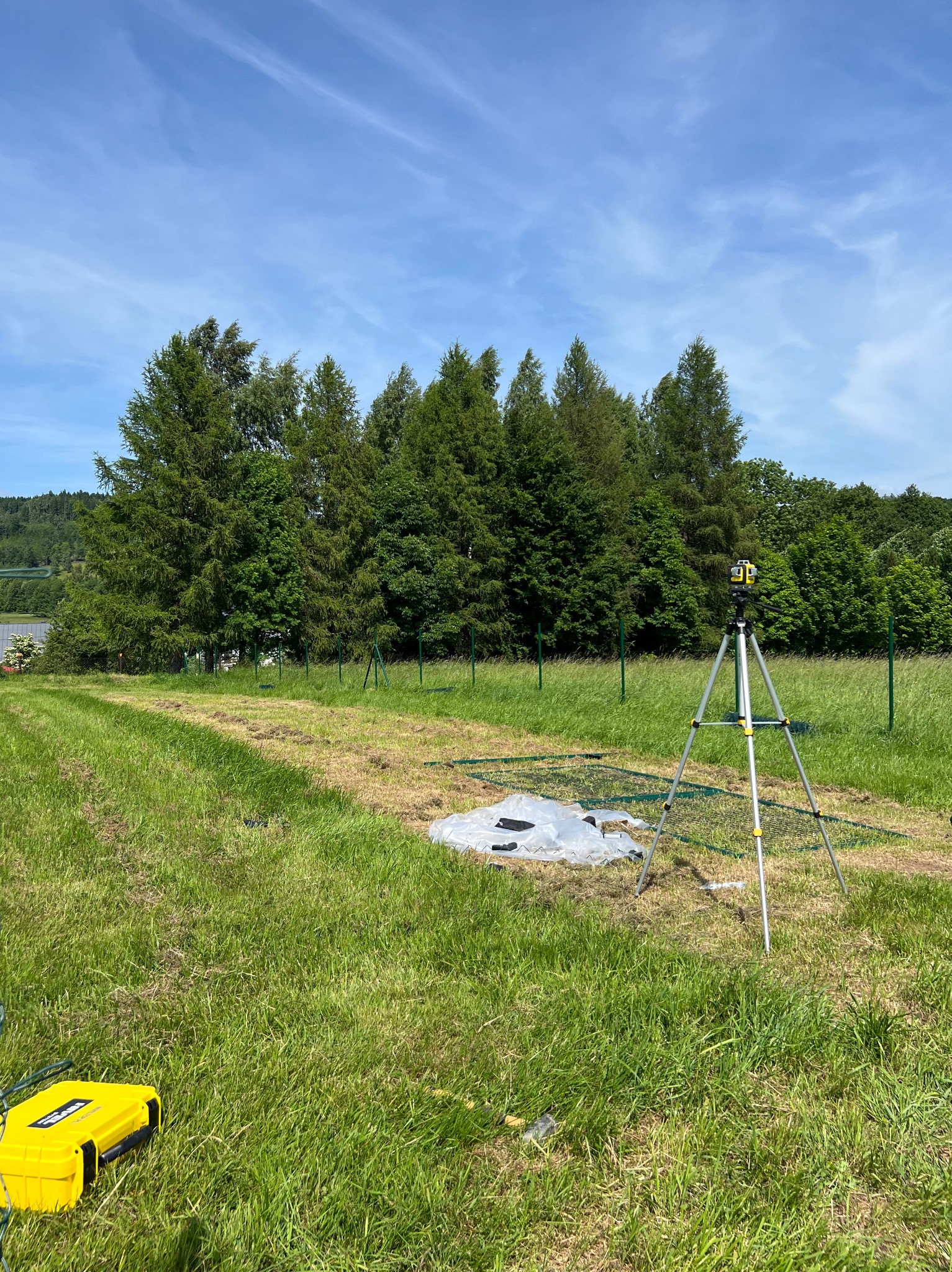 Zielony teren z rozłożoną siatką i niwelatorem na statywie. W tle widoczne drzewa i fragment ogrodzenia. Przygotowanie do montażu ogrodzenia na trawiastym terenie.