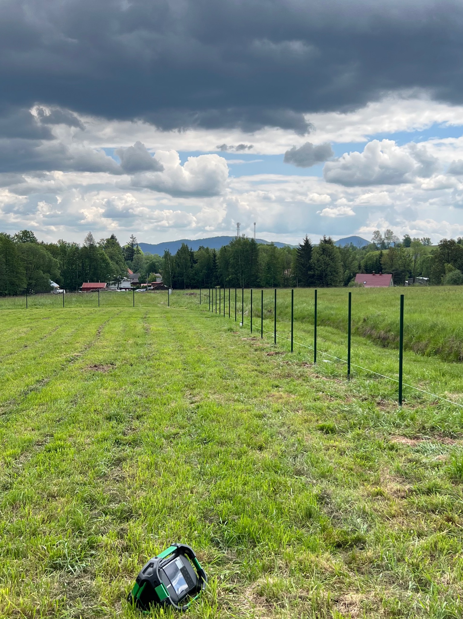 Wkopane słupki ogrodzeniowe na zielonej łące, widok na pagórkowaty krajobraz pod pochmurnym niebem. W trawie leży czarny głośnik z zielonymi akcentami.