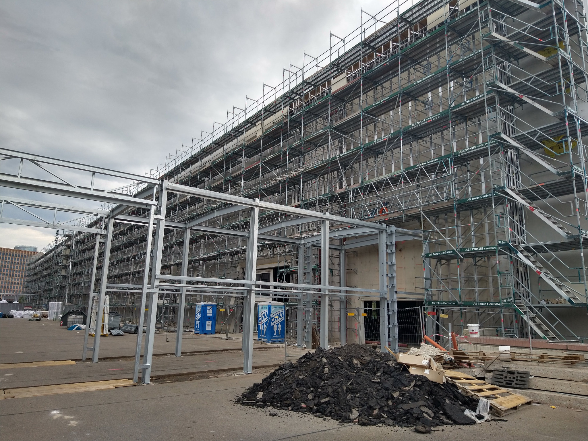 Renowacja fasady budynku w Niemczech. Rozległe rusztowania pokrywają elewację. Widoczne elementy konstrukcyjne i tymczasowe toalety. Prace w toku, teren budowy.
