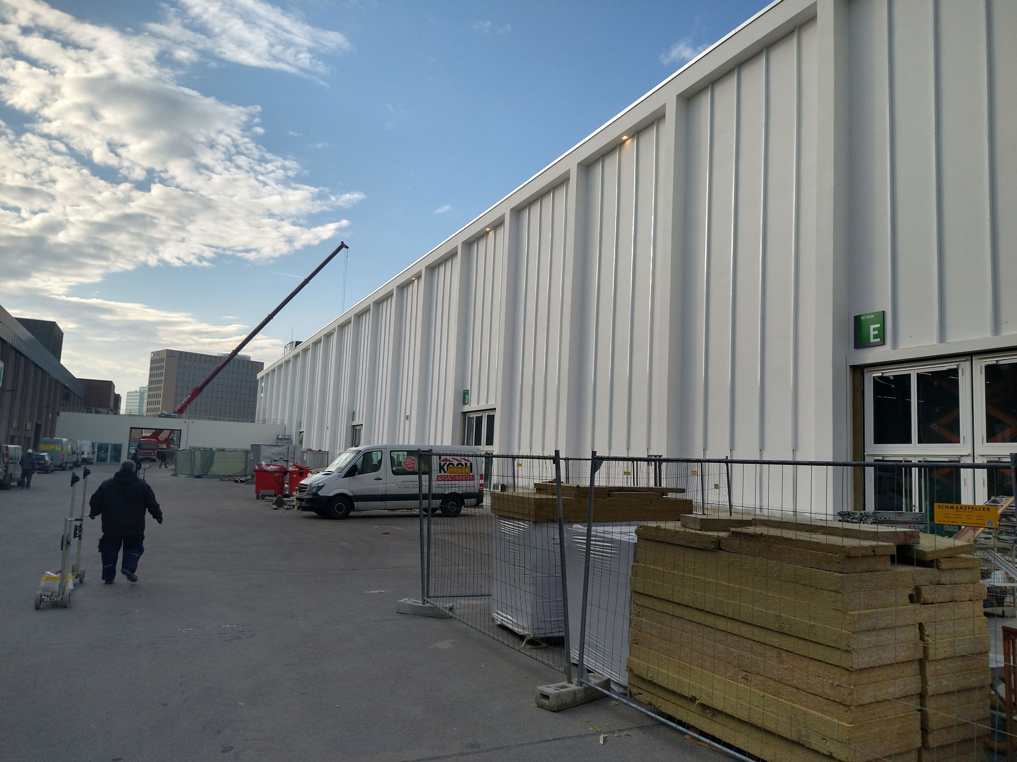 Biała hala z blachy trapezowej w trakcie budowy, widoczne materiały izolacyjne i pracownik z wózkiem. W tle dźwig i budynki miejskie pod błękitnym niebem.