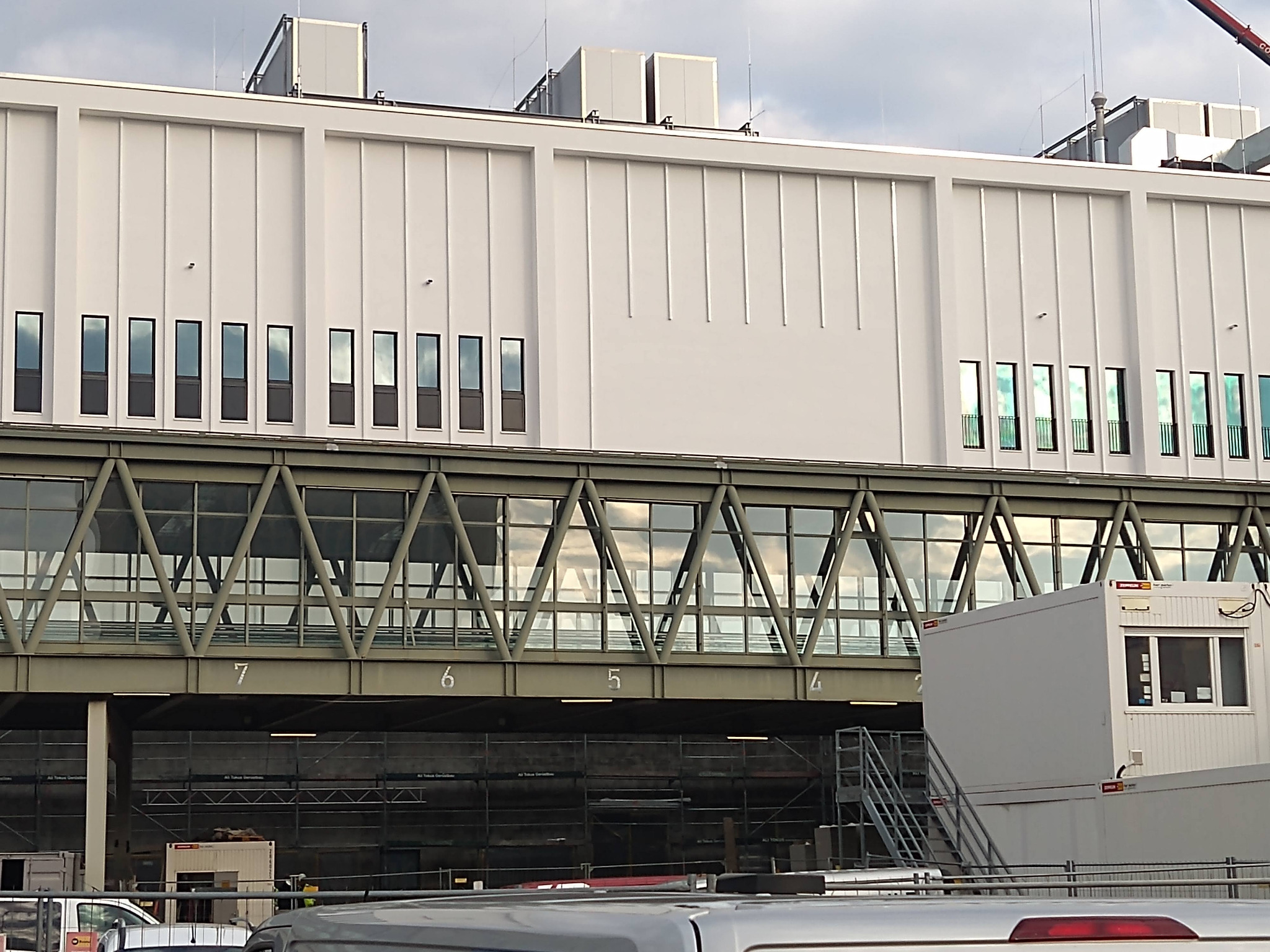 Biały budynek z przeszklonym łącznikiem nad rusztowaniami i kontenerami biurowymi, widok z dołu, częściowo zasłonięty przez dach samochodu.