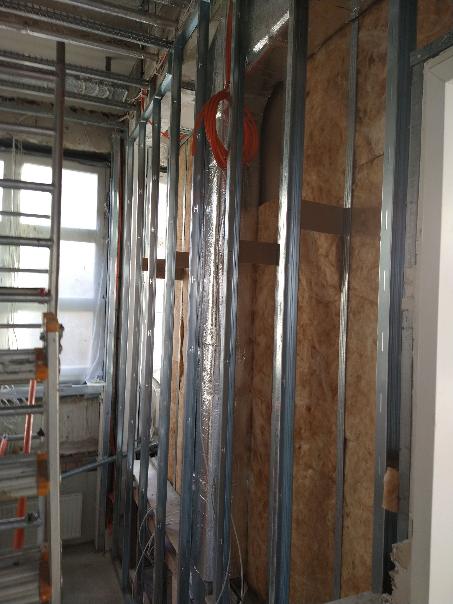 Montaż izolacji z wełny mineralnej w metalowym stelażu, widoczne przewody i rury. Prace wykończeniowe w pomieszczeniu z oknem i drabina.