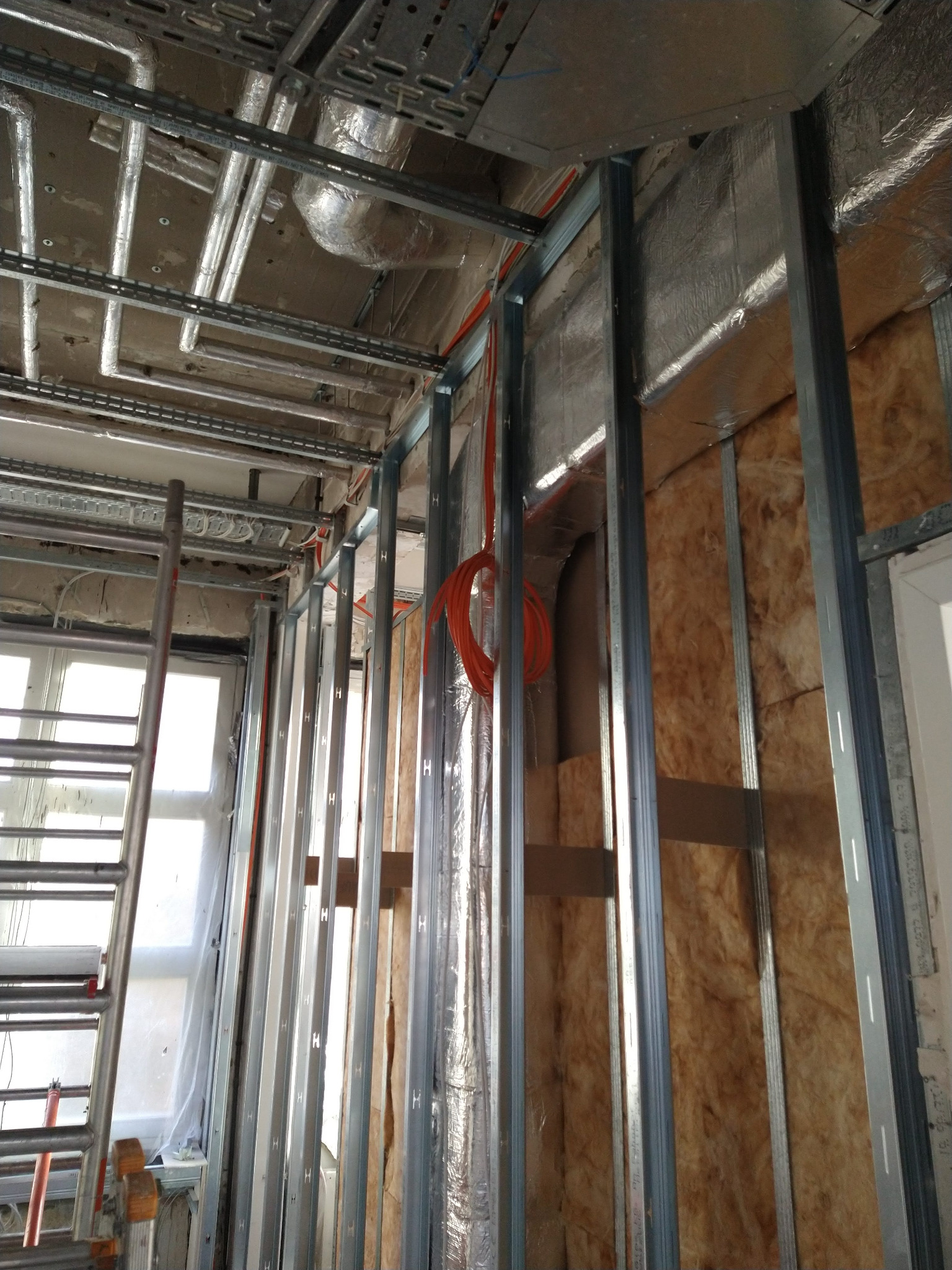 Montaż izolacji z wełny mineralnej w metalowym stelażu, widoczne przewody elektryczne i drabina. Prace wykończeniowe wewnątrz budynku.