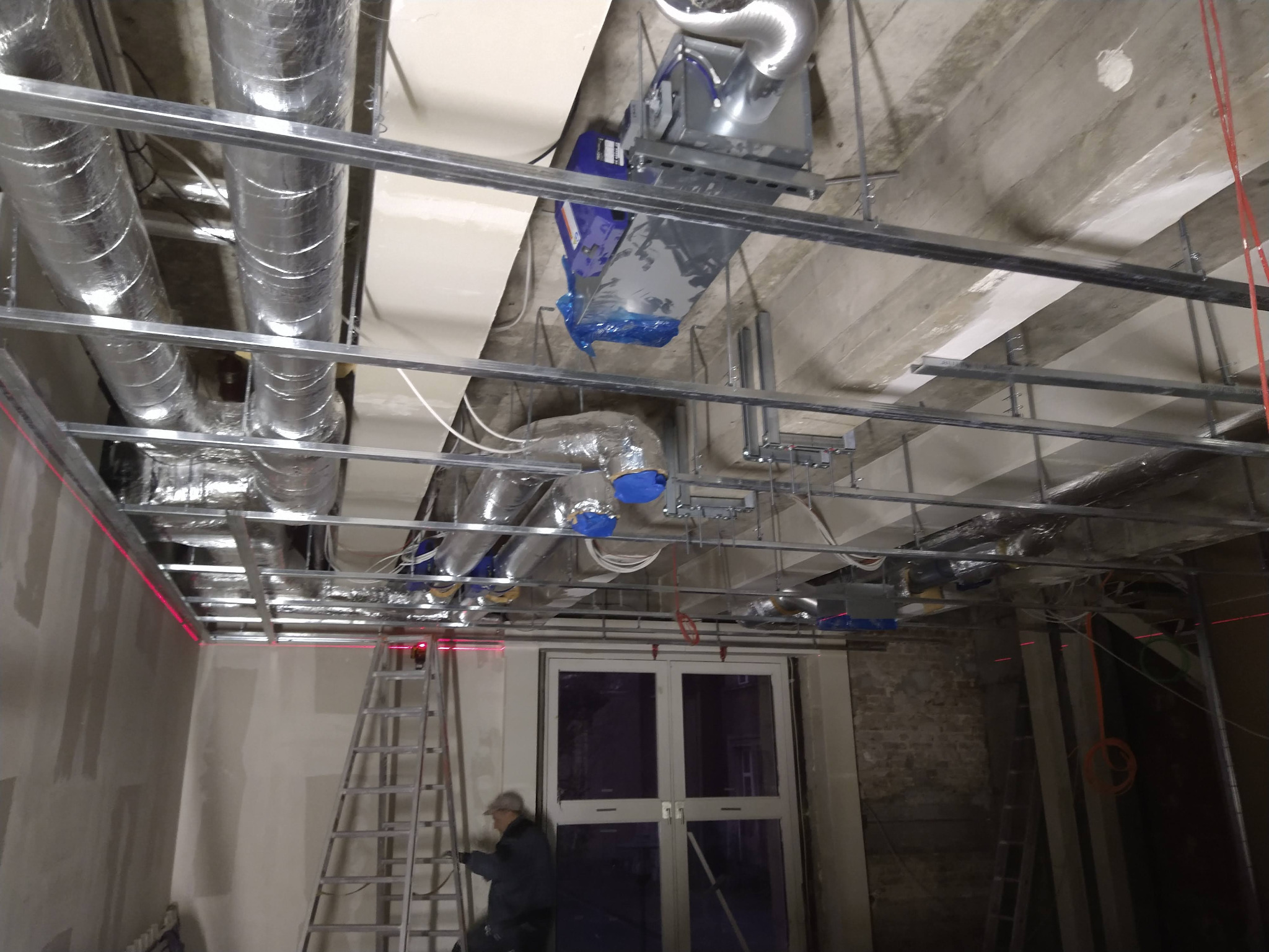 Montaż instalacji wentylacyjnej pod sufitem podwieszanym w pomieszczeniu z oknem. Widoczne rury wentylacyjne, metalowa konstrukcja i drabina. Laserowy poziom wyznacza linię.