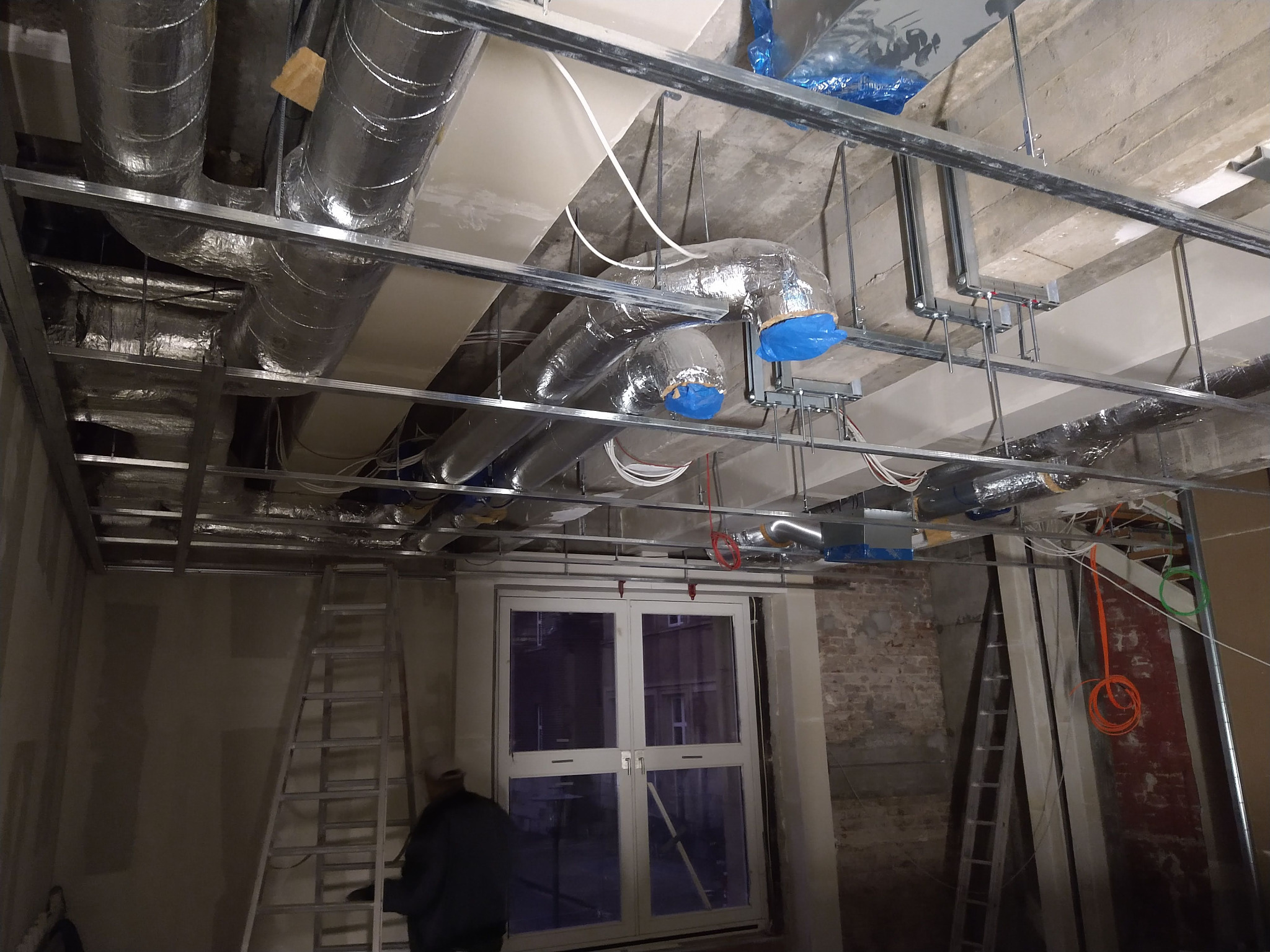 Montaż instalacji wentylacyjnej pod sufitem podwieszanym w surowym wnętrzu. Widoczne rury wentylacyjne, metalowa konstrukcja, drabina i fragment osoby.