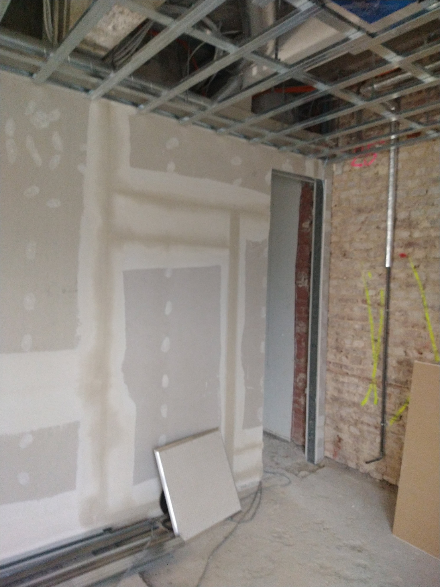 Wnętrze w trakcie prac: ściany z płyt gipsowo-kartonowych, częściowo wyszpachlowane, widoczna metalowa konstrukcja sufitu podwieszanego i fragment ściany z cegły.