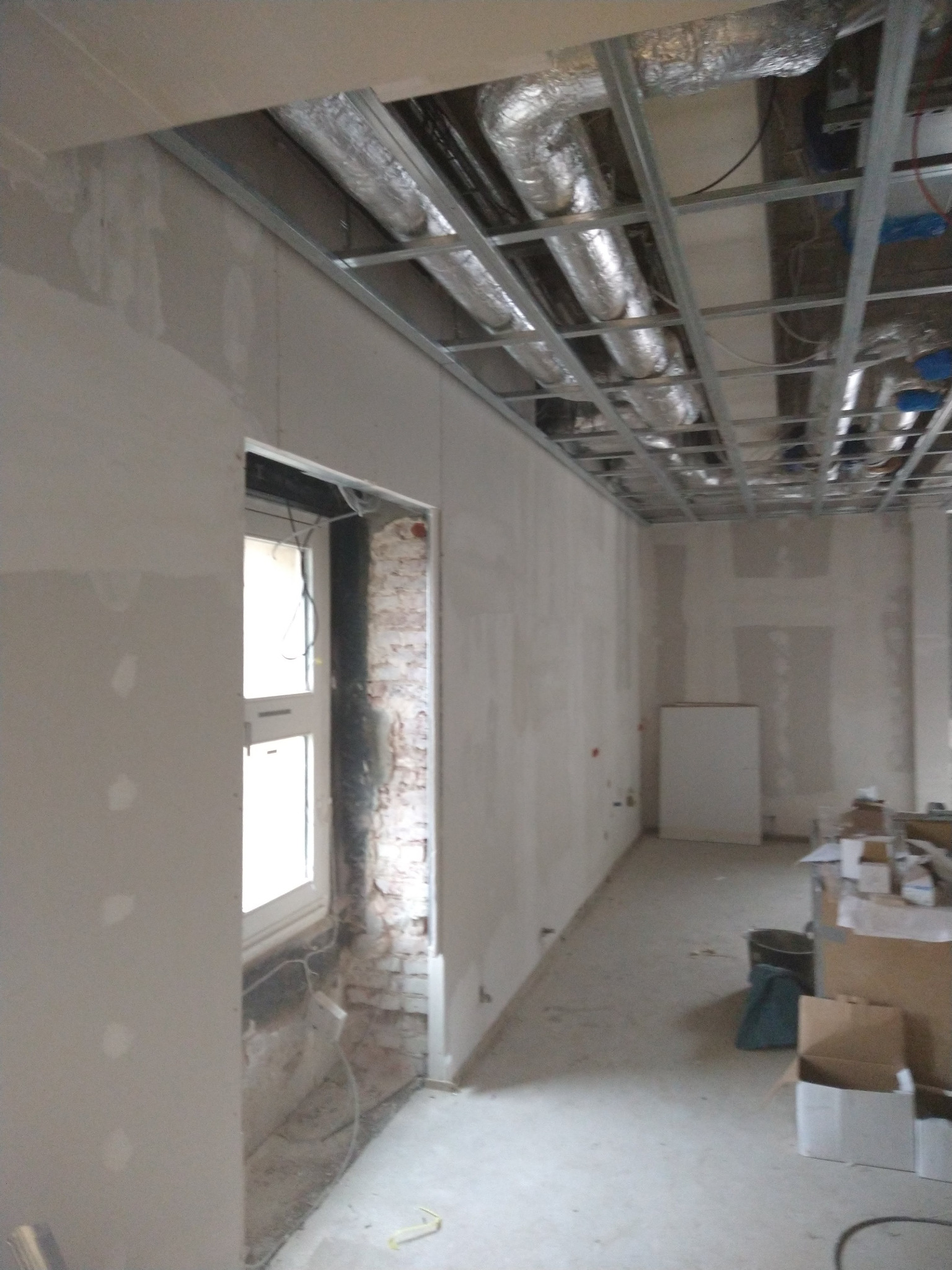 Wnętrze w trakcie remontu: ściany z płyt karton-gips, odsłonięta cegła przy oknie, metalowa konstrukcja sufitu z instalacją wentylacyjną. Widoczne narzędzia i materiały budowlane.