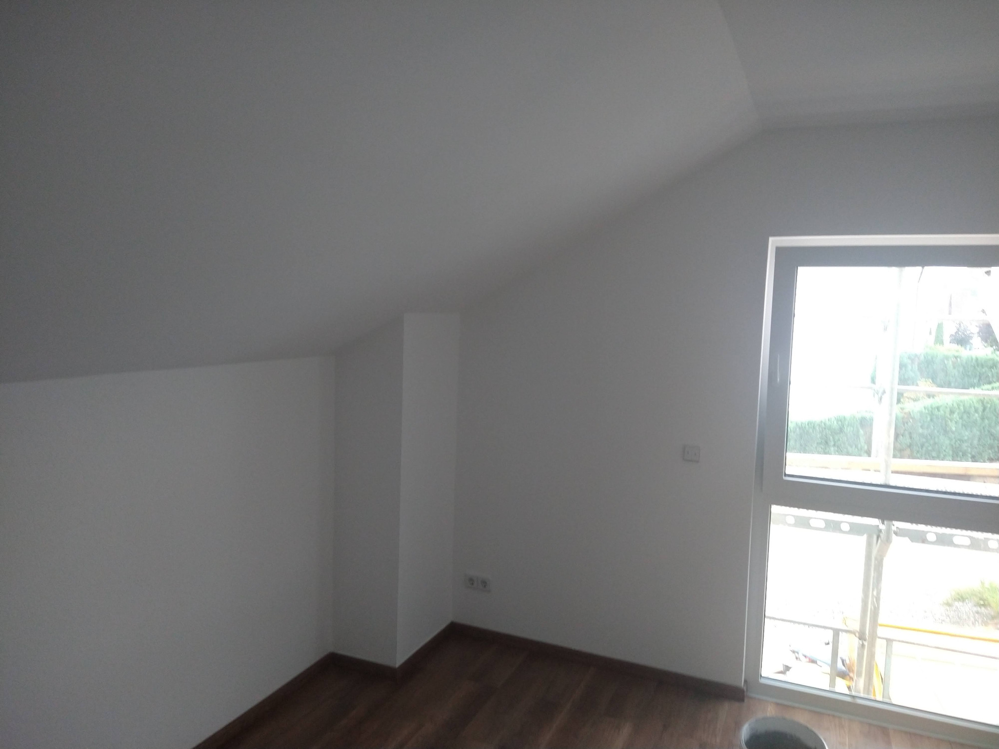 Poddasze po malowaniu na biało, widoczne drewniane panele podłogowe, okno z widokiem na zieleń i fragment rusztowania. Świeżo pomalowane ściany i sufit.