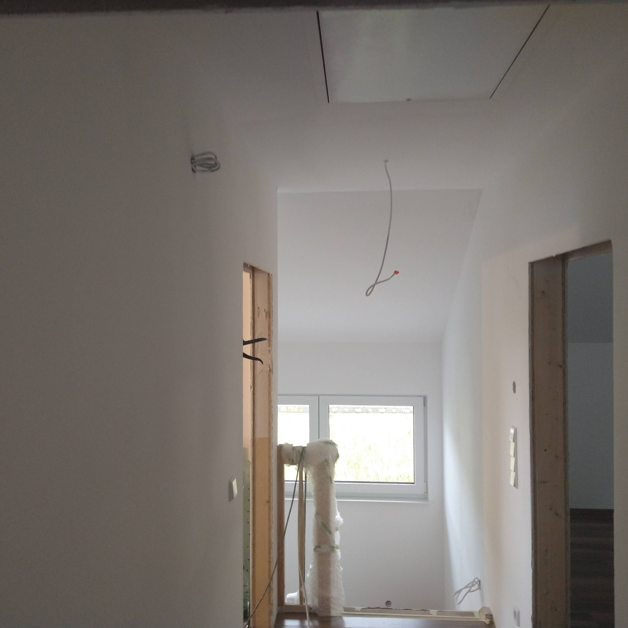 Wnętrze w trakcie prac wykończeniowych: białe ściany, sufit z otworem rewizyjnym, widoczne przewody elektryczne, ościeżnice drzwiowe i okno. Elementy zabezpieczone folią.